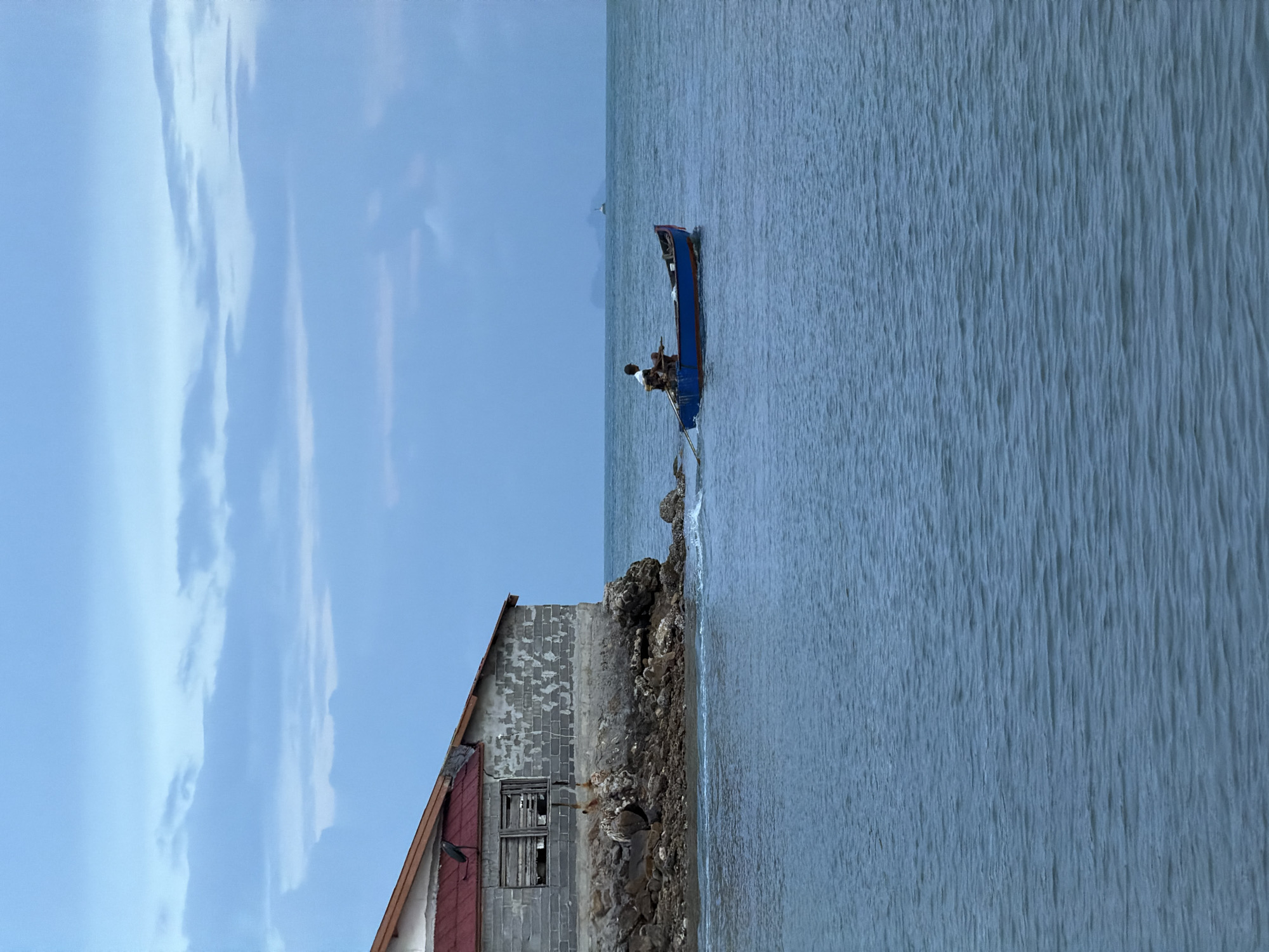 A solitary person is rowing a small blue boat in the ocean off the coast of Ko Samui District, Thailand. The boat is being pulled by a rope attached to an abandoned, weathered building on the shore. The building is made of concrete blocks and has a red tiled roof. It appears to be in disrepair, with a broken window. Rocks and debris are visible along the shore in front of the building. The ocean water is a calm, muted blue, with gentle ripples. The sky above is a soft, cloudy blue, with a hint of evening light. In the distance, a faint silhouette of land and a small structure, possibly a lighthouse, can be seen on the horizon. The overall scene evokes a sense of isolation and stillness.