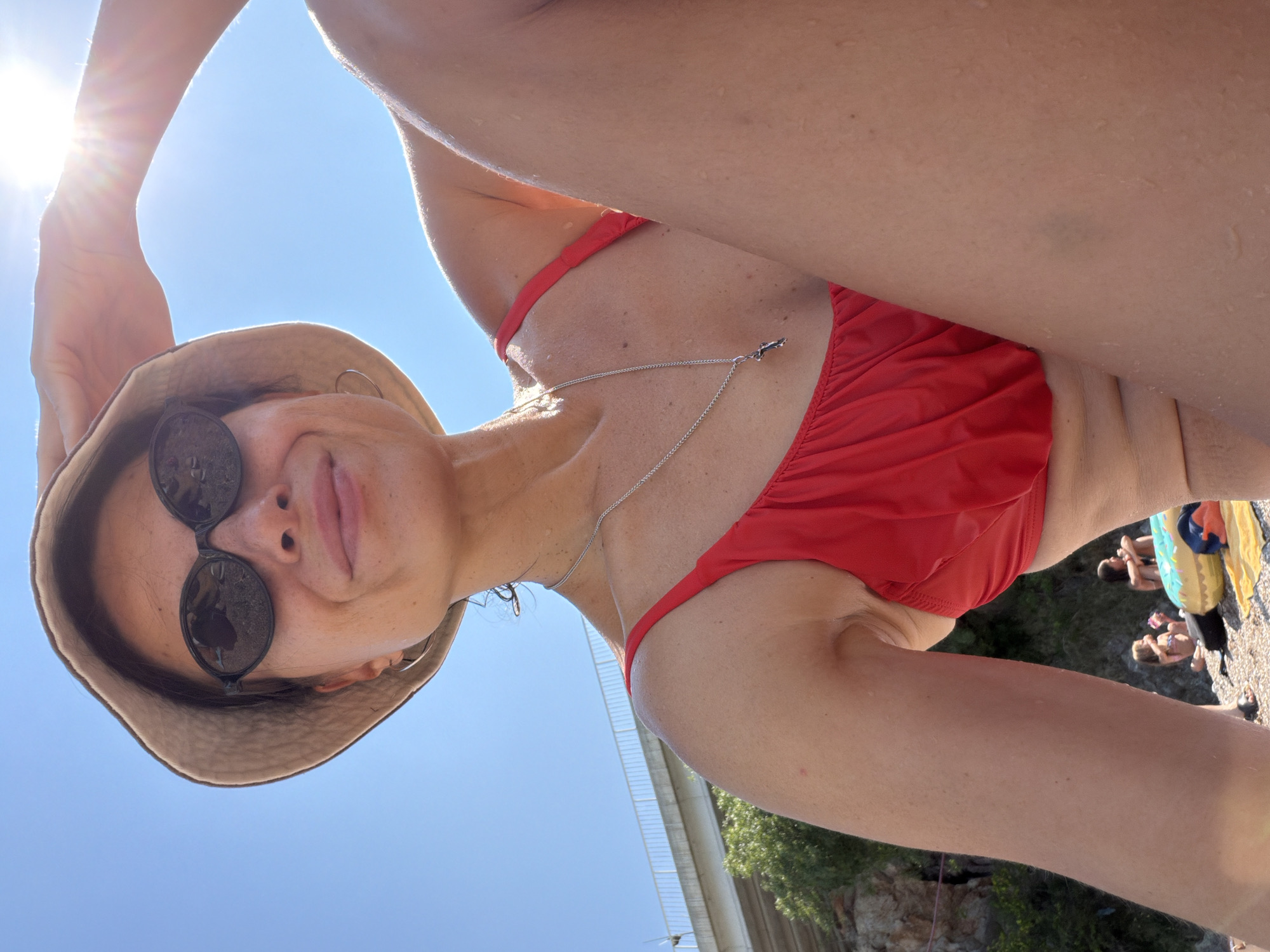 In a medium shot, a woman wearing a red bikini and a tan bucket hat poses for a selfie on a sunny beach. She is looking at the camera with a slight smile and her left hand is resting on her hat. Her sunglasses are reflecting the bright sun. A silver necklace with a cross pendant is visible around her neck. The background shows a clear blue sky, some trees, and a bridge. Further in the background, two children are seen playing near an inflatable toy. The scene appears to be an outdoor recreational area by the water.