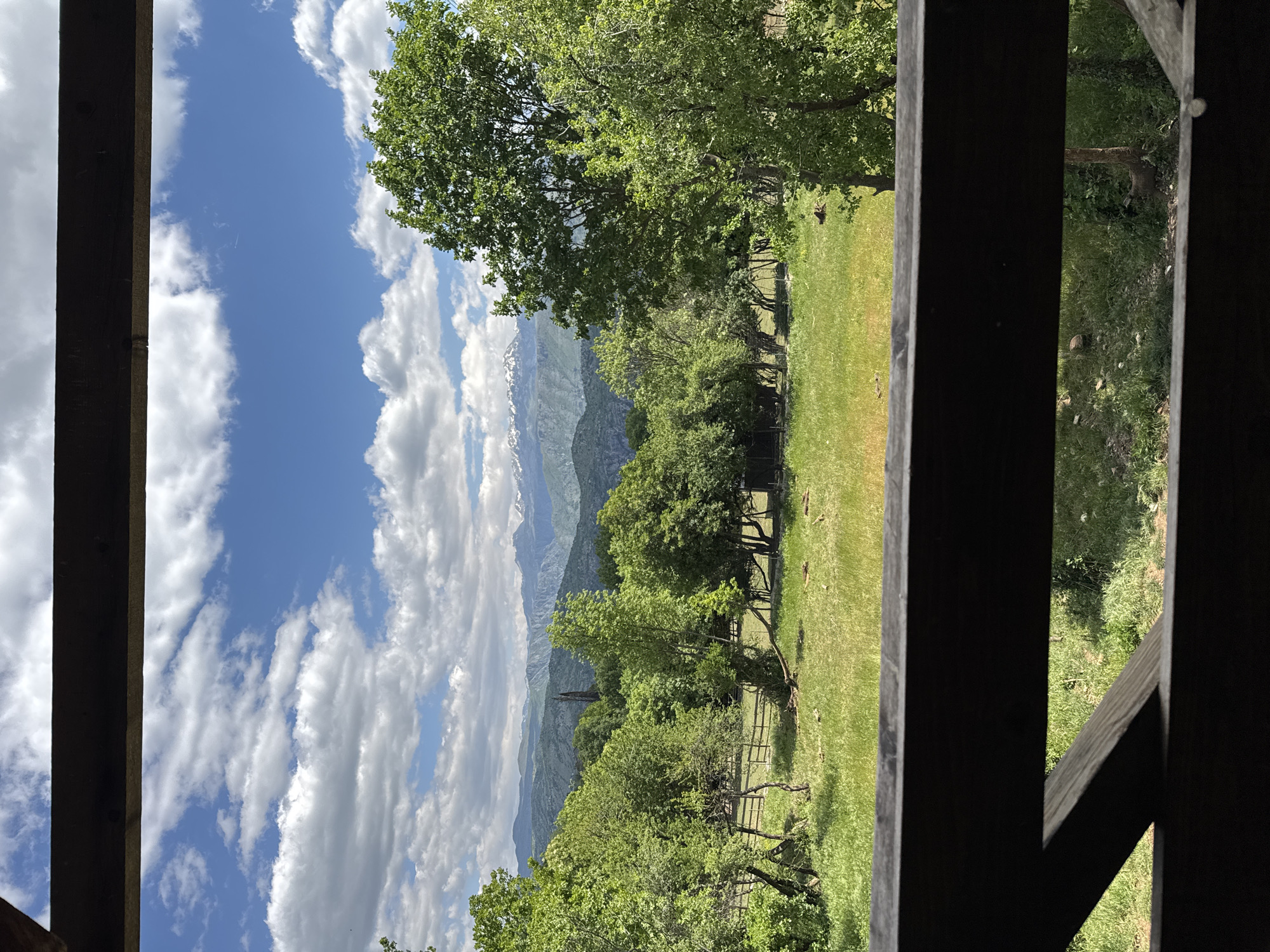 The image captures a view from within a wooden structure, likely a porch or balcony, in Gornje Mrke, Montenegro. The vantage point offers a glimpse of a mountainous landscape with snow-capped peaks partially obscured by cumulus clouds. The immediate foreground features lush greenery, including trees of varying sizes and a grassy field enclosed by a wooden fence. Dark wooden beams and supports frame the view, indicating the observer's position within a building. The sky is blue with scattered white clouds.