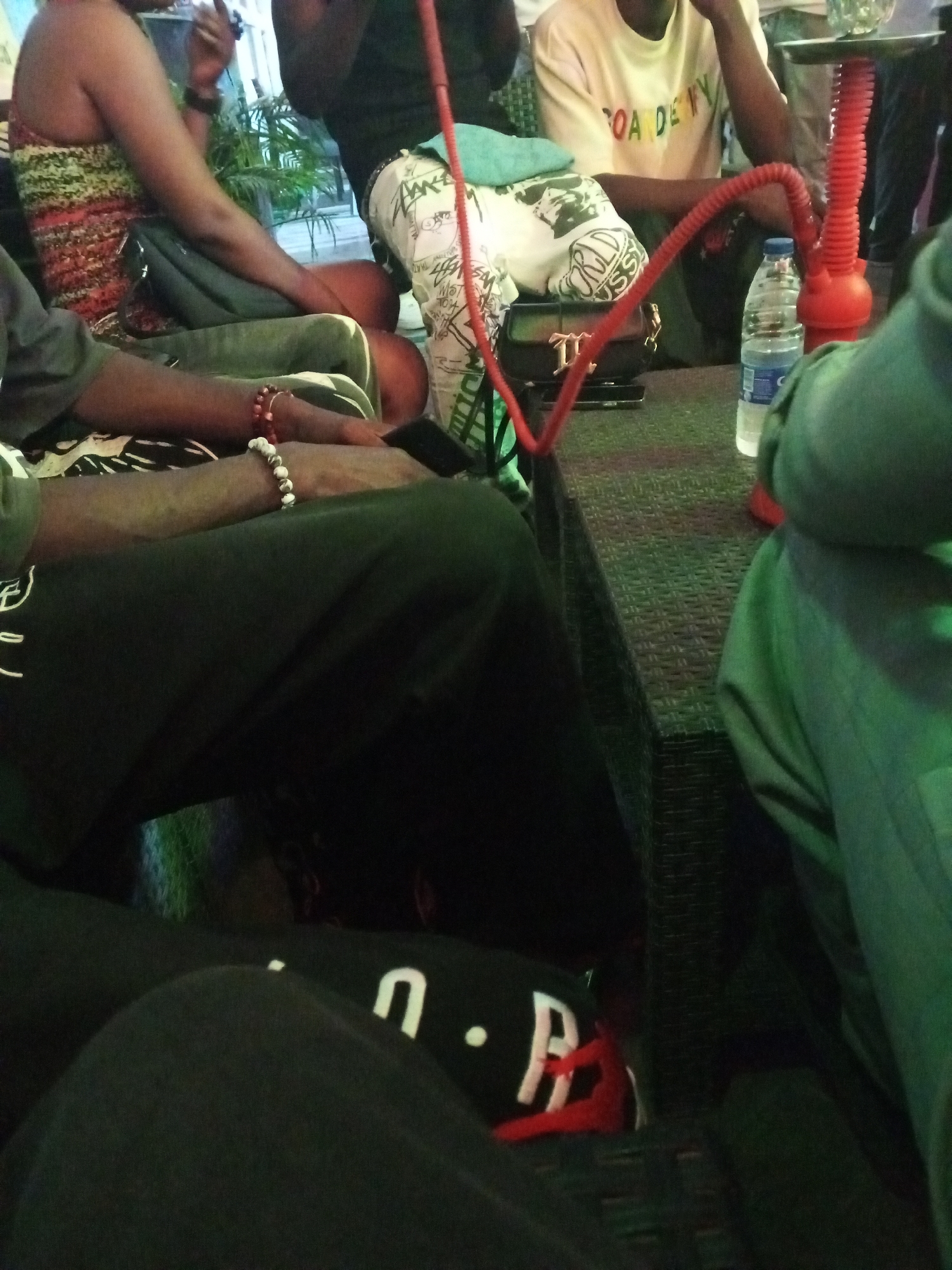 Several people are gathered around a table in what appears to be an outdoor or semi-outdoor setting in Lokoja, Nigeria. A red hookah is present on the table, along with a bottle of water. The table is textured with a woven or patterned surface. At least one person is wearing a t-shirt, while another is wearing patterned pants and a tank top. Bracelets are visible on the wrist of one of the people. A purse is situated on the table, beside the hookah. Some of the individuals are seated, while others are partially visible, cropped out of the frame.