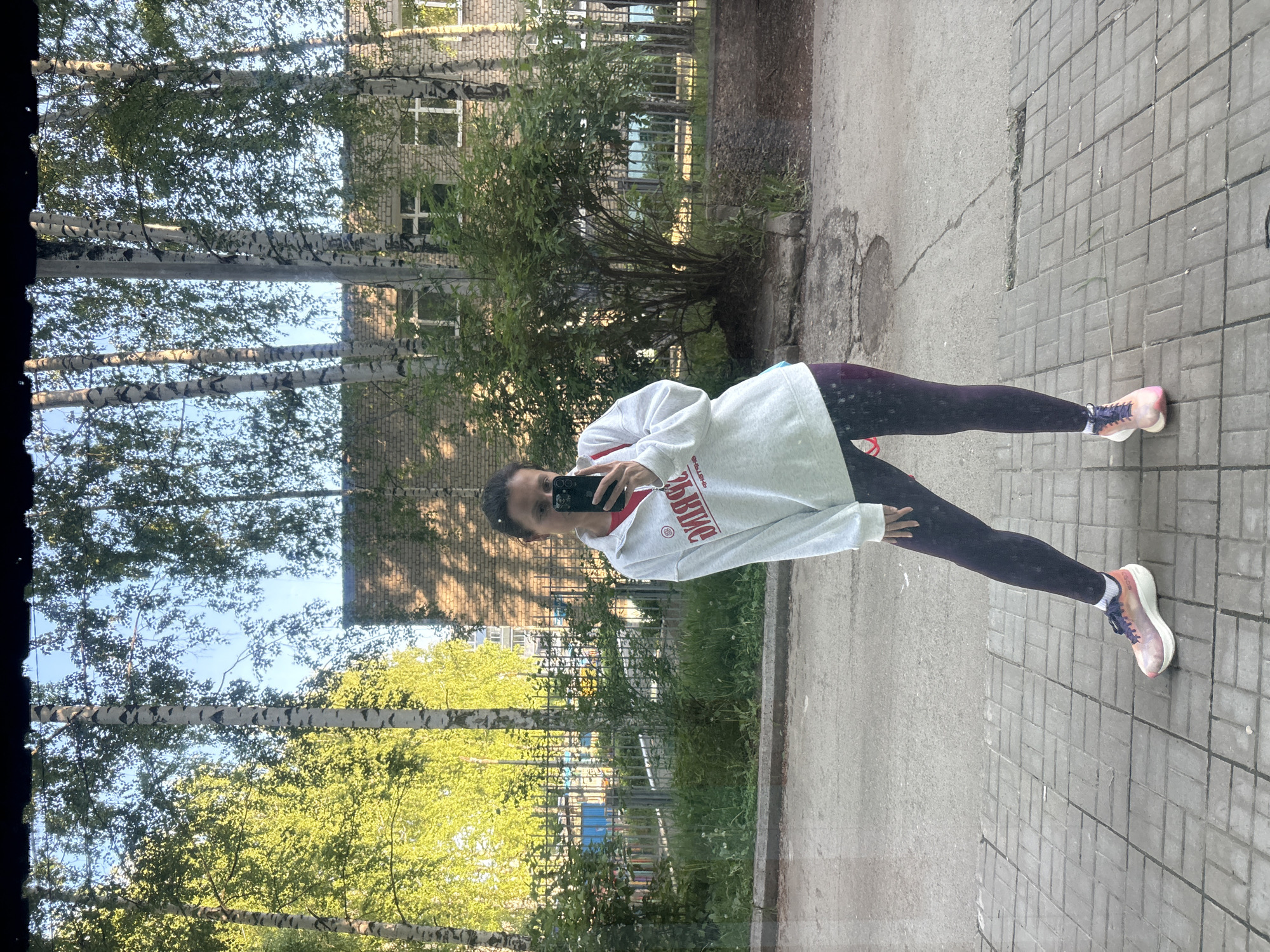 A mirror selfie captures a person standing outdoors in what appears to be a residential or park area in Chaikovsky, Russia. The image is a reflection, showing the person holding a black smartphone, likely an iPhone, to take the picture. The individual is wearing a light grey long-sleeved top, possibly a hoodie or sweatshirt, with dark leggings and multi-colored athletic shoes featuring pink, purple, and white accents.
The background reflected in the mirror depicts a bright, sunny day. Several tall birch trees with light green leaves are prominent, indicating springtime or early summer. Behind the trees, a light-colored brick building with multiple windows is visible, along with a dark metal fence and some green bushes. The ground consists of a paved sidewalk made of grey rectangular tiles and an asphalt path with manhole covers. The overall setting suggests a typical urban street or pedestrian area.
The person’s face is partially obscured by the smartphone, making specific emotions hard to discern, but the pose is casual. On the person's grey top, red text is visible, which appears to say "ЯВЛЕНИЕ" (YAVLENIYE), meaning "PHENOMENON" or "APPEARANCE" in Russian. Some other less clear Russian text is also present. The bright daylight and lack of harsh shadows suggest it is either morning or late afternoon.