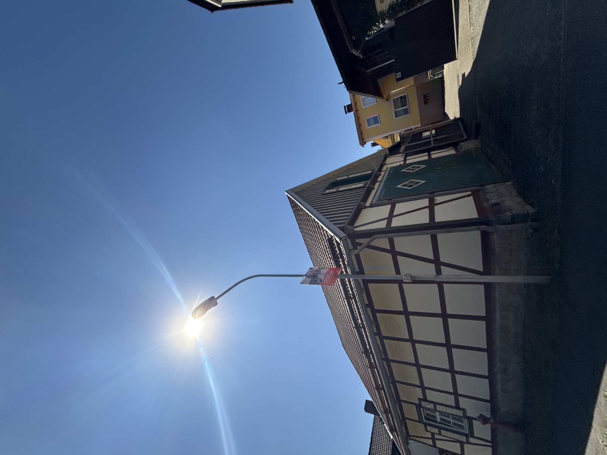 In Bad Sachsa, Germany, a timber-framed building with a red-tiled roof is depicted under a clear blue sky and bright sunlight. A lamppost stands in front of the building, adorned with a political poster featuring a man's face and text. Adjacent to the timber-framed structure, a yellow house and a dark building are visible, suggesting a street or alleyway. The scene is bathed in sunlight, casting shadows on the ground and the buildings.