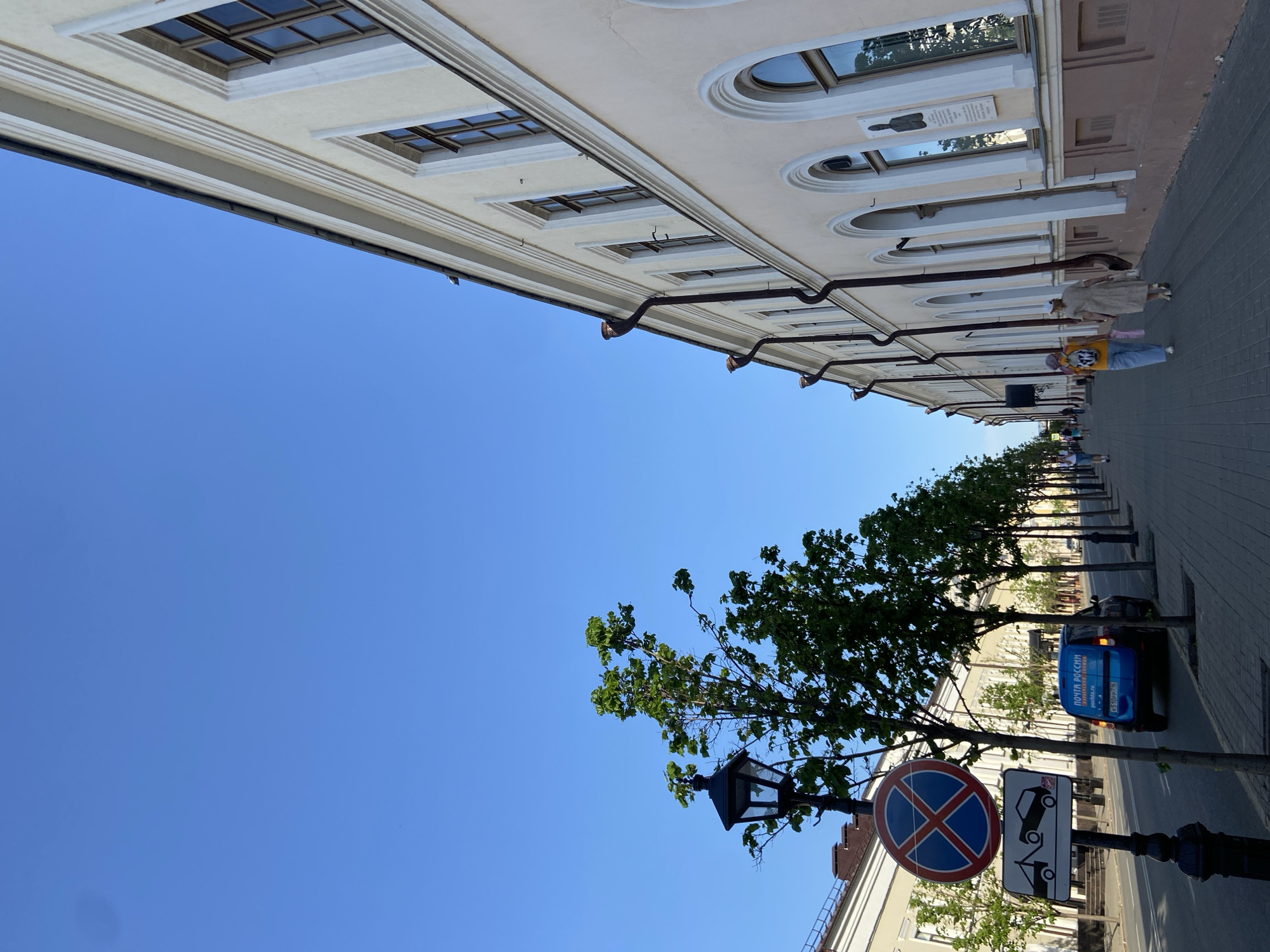 The image shows a street view in Kazan, Russia. A light-colored building with multiple windows runs along the top edge of the frame. Below it, a line of trees with green leaves stretches along the side of the street. A "No Parking" sign is visible near a lamppost in the foreground. A blue delivery van, possibly for "ПОЧТА РОССИИ" (Russian Post), is parked next to the curb. Two people are walking on the sidewalk. The sky is clear and blue. The overall composition is a low-angle shot, emphasizing the height of the building and the open sky.