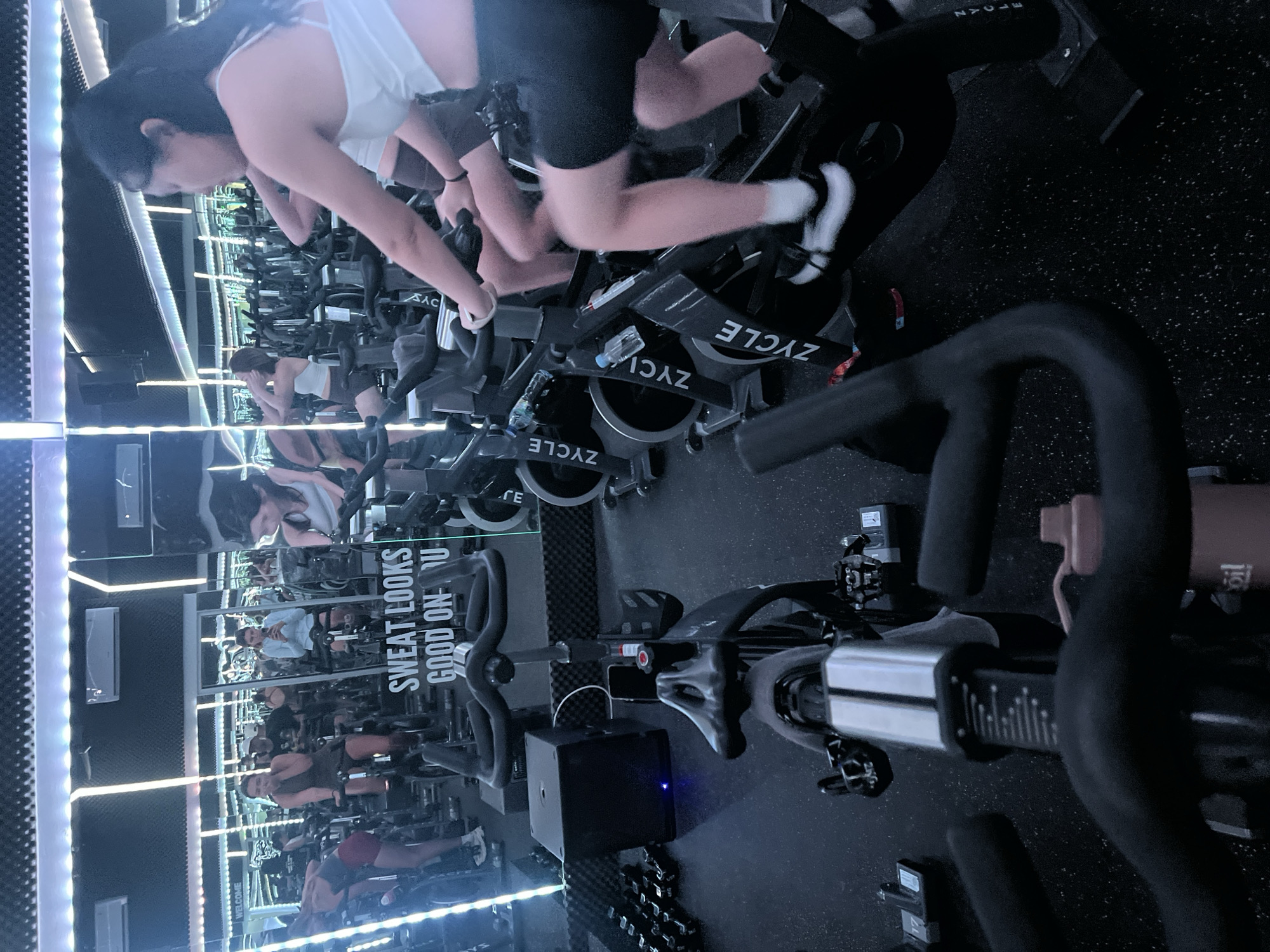 A group of people are engaged in a spin class at a fitness studio, likely in North Kuta, Indonesia, based on the provided information. The room is dimly lit with accent lighting along the ceiling and walls, and mirrors line the back wall, reflecting the scene. In the foreground, a black spin bike is positioned centrally, with its handlebars and seat clearly visible. To the right and slightly behind, another spin bike is occupied by a woman with long dark hair wearing a white tank top and black shorts, actively pedaling. Several other spin bikes and participants are visible throughout the studio, some in the foreground and others receding into the background, all engaged in the exercise class. Some individuals are in focus, while others appear blurred due to motion. The floor is a dark, speckled rubber material typical of gym environments. A sign with the text "SWEAT LOOKS GOOD ON U" is visible on the mirrored wall. The overall atmosphere suggests a high-energy fitness session.