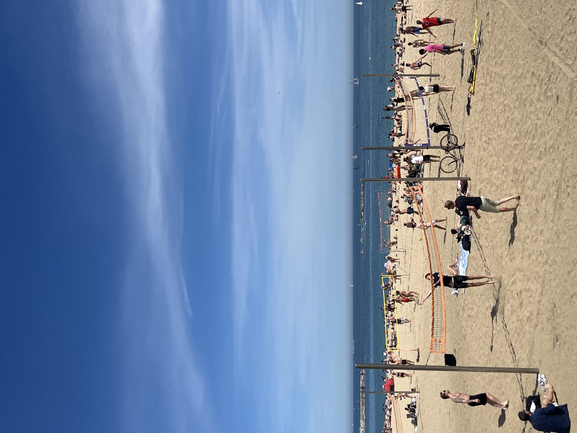 The image depicts a sunny day at Barceloneta Beach in Barcelona, Spain. The scene is split between a clear blue sky with some light clouds and a crowded sandy beach. The Mediterranean Sea is visible in the background, with several boats sailing on the water.
The beach is populated with people sunbathing, playing volleyball, and relaxing. Several volleyball nets are set up, with people actively playing games. Other people are walking, standing, or sitting, scattered across the sand. There are bicycles parked near the volleyball nets.