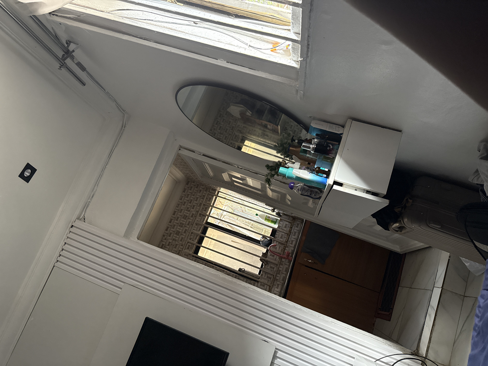 The image depicts an indoor scene, possibly a bathroom or dressing area, within an apartment in Abuja, Nigeria. The room is lit by natural light streaming in from a window. A white-tiled floor is visible in the foreground, and an open doorway reveals a glimpse of a kitchen area with wooden cabinets and a window secured by metal bars.
On the right side of the frame, a large oval mirror is mounted on the wall, reflecting part of the room. Below the mirror, a white shelf unit holds various cosmetic products, including bottles of lotions and perfumes, as well as a small decorative Christmas tree. To the left of the mirror, a white vertical structure with ribbed detailing lines the wall.
A dark-colored suitcase is positioned on the floor near the doorway. A black television screen is partially visible on the far left of the image. The walls are painted white, and the ceiling has recessed lighting. Electrical cords are neatly organized along the ceiling and wall. The overall impression is of a lived-in space.