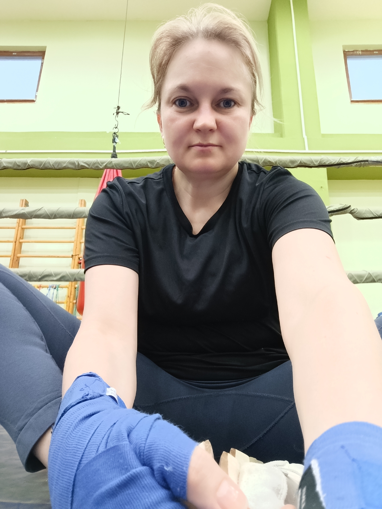 A woman with blonde hair sits in what appears to be a boxing ring. She is wearing a black T-shirt and dark leggings. Her hands are wrapped in blue athletic tape. The ring is surrounded by green walls, with a pair of small windows visible in the background. There is a ladder-like structure behind the ring.
The city and location are unknown.