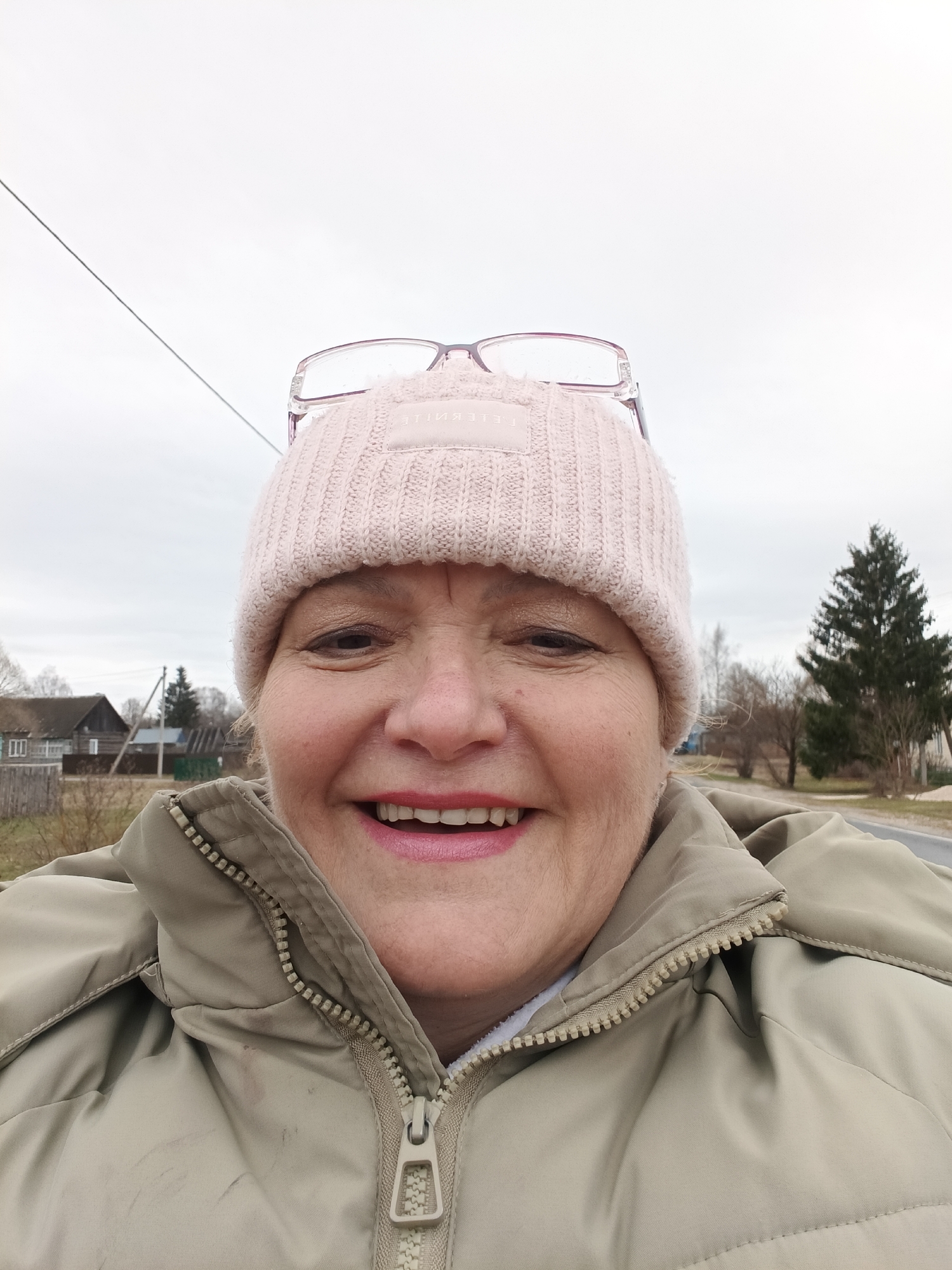 A close-up selfie shows a woman smiling with pink lipstick, wearing a light pink knit hat with glasses perched on top, and a light brown padded jacket. The jacket's zipper is fully zipped. The background reveals a rural setting in daylight with overcast skies. In the distance are houses, a power line, and trees, some evergreen. The location is likely within Красный Маяк, Russia, judging from the architecture and landscape.