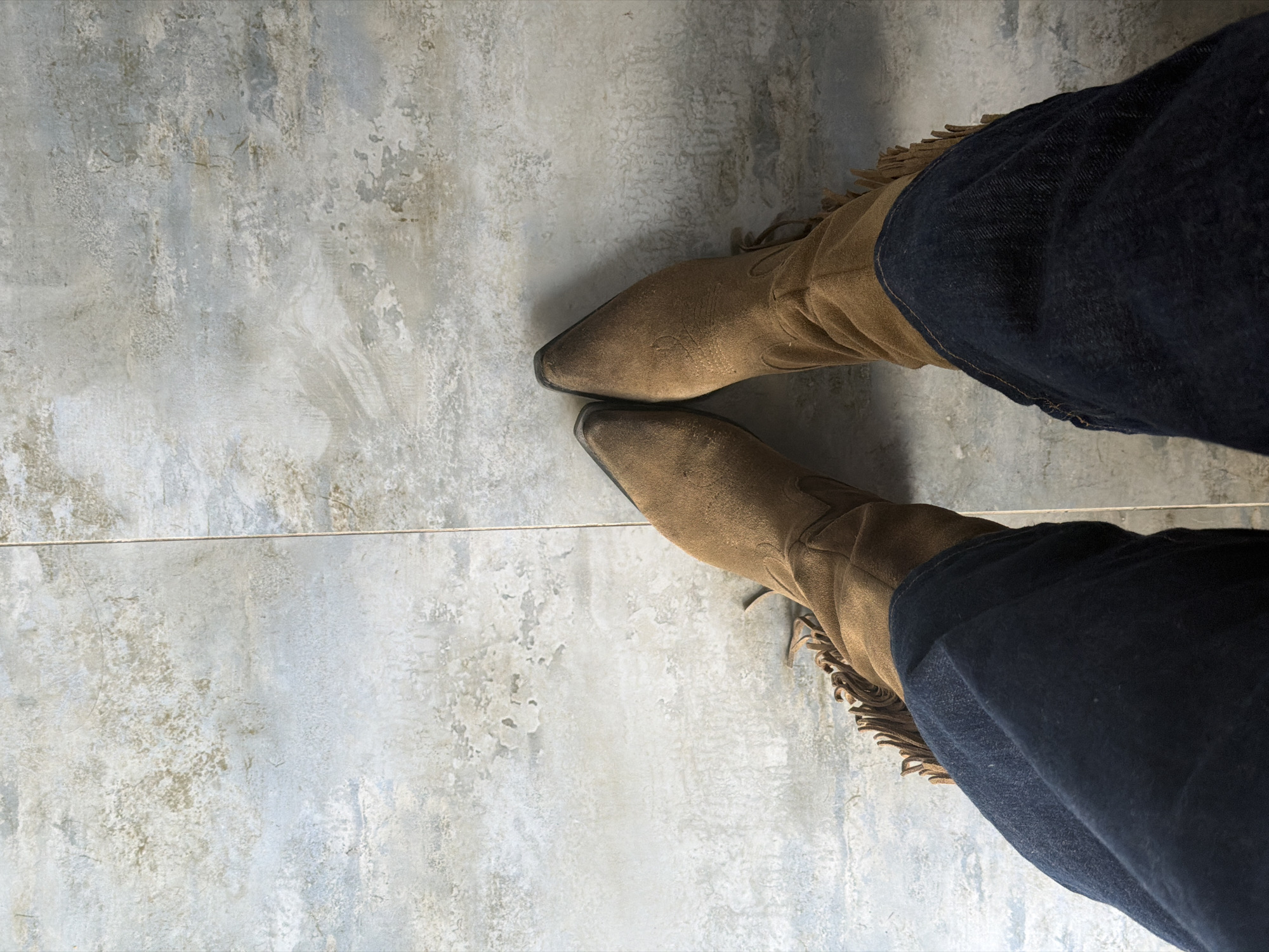 The image captures an overhead, slightly angled view of a person's lower legs and feet, standing on a textured light gray tiled floor. The person is wearing dark blue jeans and brown suede cowboy boots with pointed toes. The boots feature distinct fringe detailing along their outer sides. The jeans are visibly tucked into the shafts of the boots.
The floor is composed of large, rectangular tiles that exhibit a mottled, distressed pattern, resembling aged concrete or stone with variations in light gray and subtle beige hues. A thin, dark grout line runs horizontally across the mid-section of the frame, marking the boundary between two tiles. The perspective suggests the person is stationary. The setting is likely indoors in Yerevan, Armenia, given the tiled floor and even lighting. No specific activity or event is taking place beyond standing. There is no visible text, and emotions or specific time of day are not discernible.