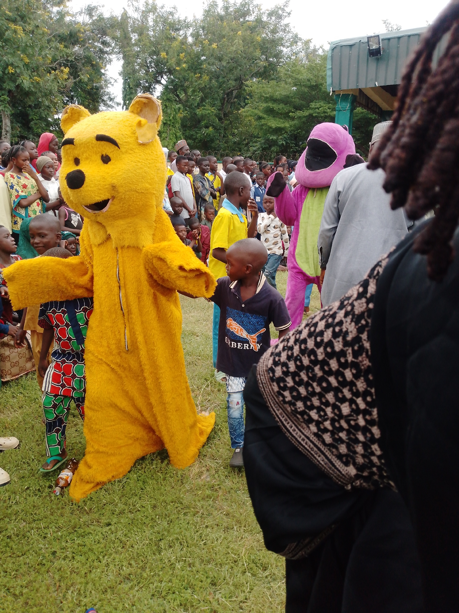 A group of children and adults are gathered on a grassy field in front of a covered structure with a metal roof. A person in a full yellow Winnie the Pooh costume stands in the foreground, facing left and gesturing with an open hand. To the right of Winnie the Pooh, a child wearing a dark t-shirt with a "Burberry" logo looks away from the camera. Behind Winnie the Pooh, a person in a pink dinosaur costume is visible. The background is filled with trees and more people. A bottle of what appears to be malt beverage is lying on the grass near the feet of Winnie the Pooh.