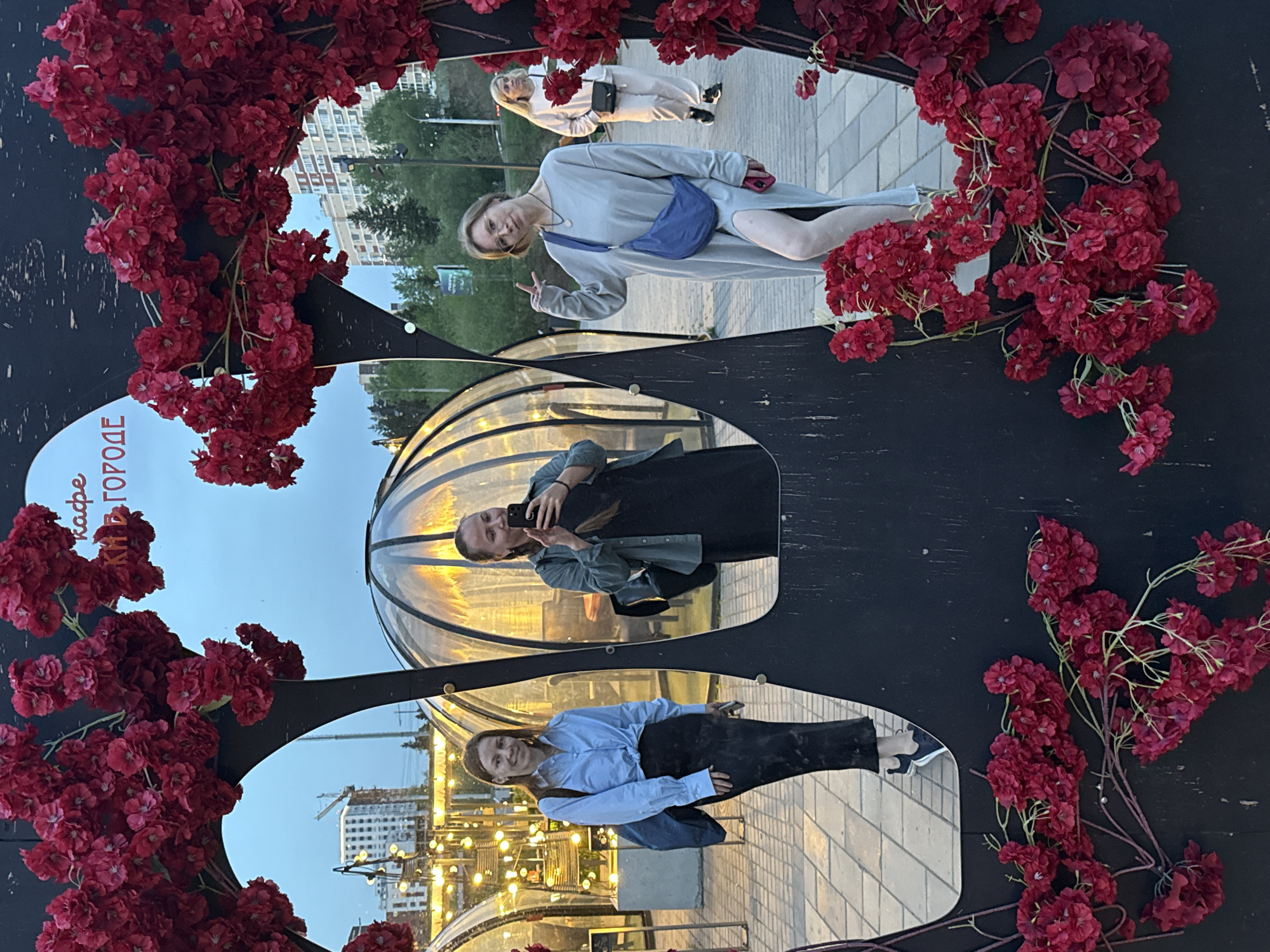 The image shows three women reflected in a mirrored public art installation in Izhevsk, Russia. The installation is decorated with red flowers. The top woman is wearing a gray dress and a blue bag, and she is making a peace sign. Another woman, standing behind her, is wearing a white outfit. The second woman in the middle mirror is wearing a black dress and a green jacket, and she is holding a cell phone to take a picture. The third woman in the bottom mirror is wearing a blue shirt and a black skirt. In the background are buildings and streetlights.