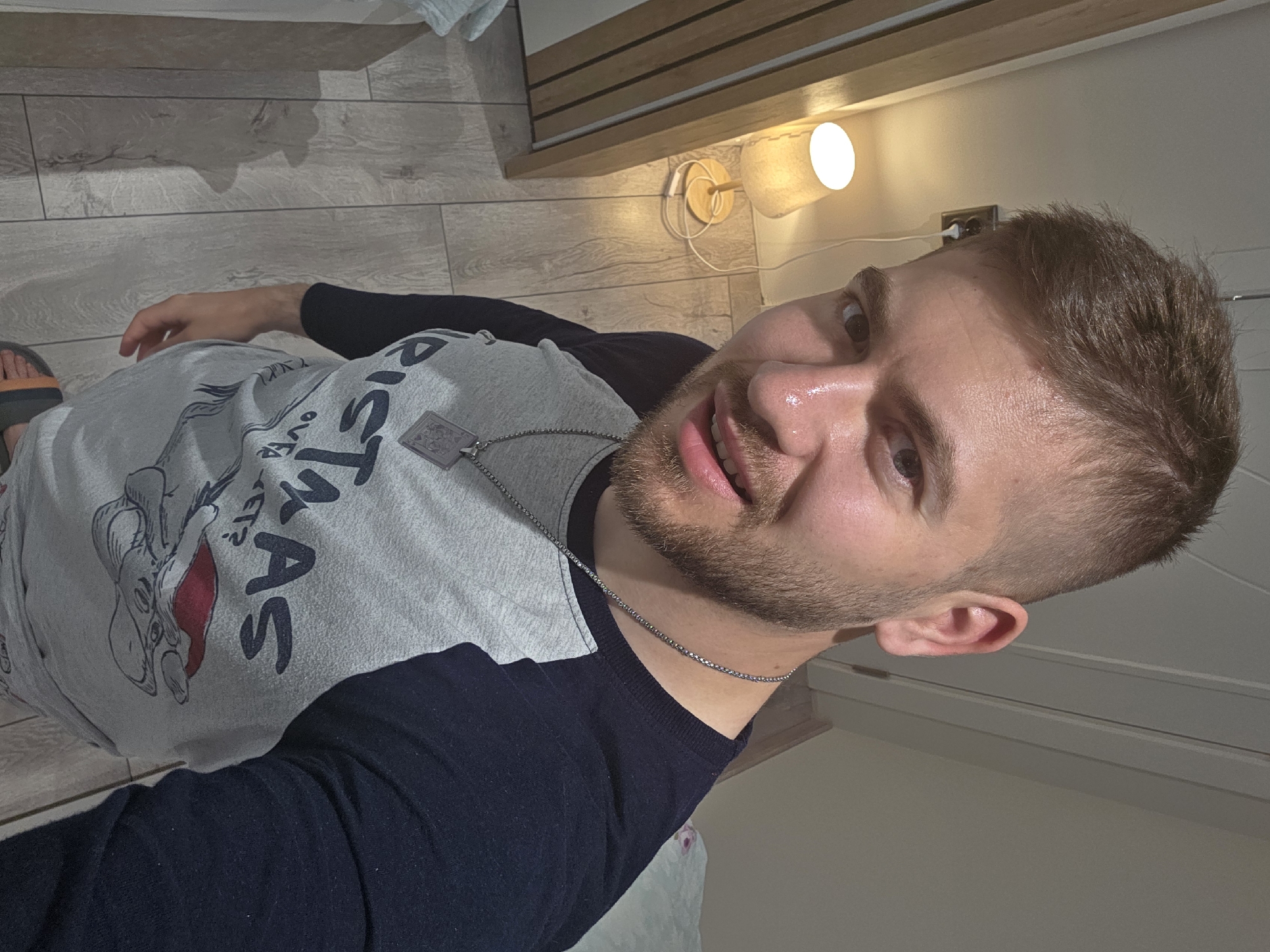The photograph shows a man with short, light brown hair and a trimmed beard, wearing a gray t-shirt with a black long-sleeved under shirt and a necklace with a pendant. He appears to be indoors, possibly in a bedroom, as suggested by the presence of a wooden bed frame, a wall-mounted lamp, and wood-look flooring. The exact location within the city of Yagodovo, Bulgaria, cannot be determined from the provided image.