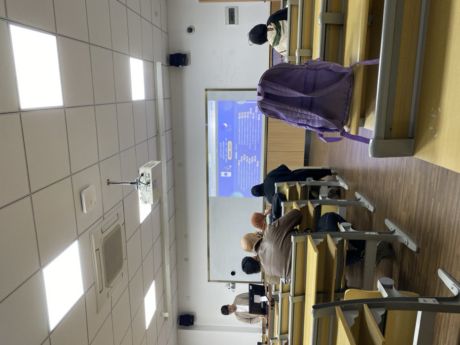 The image depicts an indoor classroom within Yinzhou District, China. The specific building or institution is not identifiable. The room features white walls, a tiled ceiling with active square LED lighting panels and a central air conditioning unit, and a wood-grain patterned floor.
At the front of the classroom, a male individual wearing a light brown cardigan and glasses stands behind a black lectern, operating a silver laptop. He is positioned to the left of a large white whiteboard, which functions as a projection screen for a ceiling-mounted white projector. The projector displays a blue-toned digital interface with text, including Chinese characters and phrases such as "NO.1 BEST APP," onto the whiteboard. Black speakers are mounted on the side walls near the front, and a dome-shaped security camera is visible on the right wall.
Approximately five individuals are seated in student desks, which are composed of light wood surfaces and grey metal frames, arranged in rows. A student in a brown leather jacket is visible in the middle foreground, seated with their back to the camera. To their left, another student wears a light-colored headscarf. Further back, an individual in a black full-length garment is seated. On the far right, a student wearing a patterned head covering is visible from behind. No direct interactions among the individuals are apparent.
In the immediate right foreground, a purple backpack rests upright on an empty student desk, facing away from the camera. The classroom appears brightly lit and orderly.