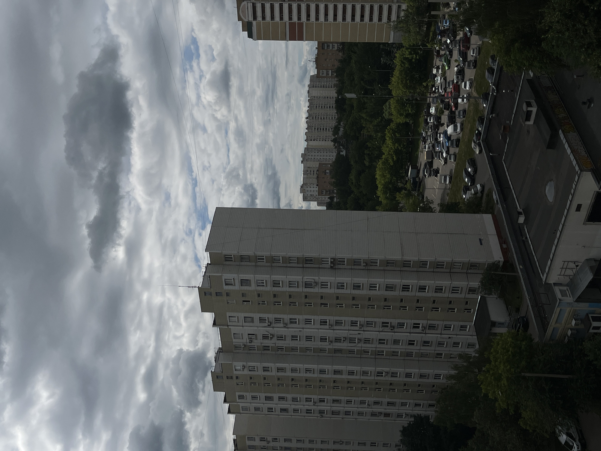 The image depicts a view of a residential area in Moscow, Russia, characterized by Soviet-era apartment buildings and a cloudy sky. In the foreground, a tall, light-colored apartment building with numerous windows is prominent. To its right, another similar building is partially visible. Further in the background, a long, lower apartment complex stretches across the horizon, nestled amongst a dense canopy of green trees. A large parking lot filled with various cars is situated in front of the distant buildings. The overall scene suggests a typical urban landscape under overcast weather conditions.
