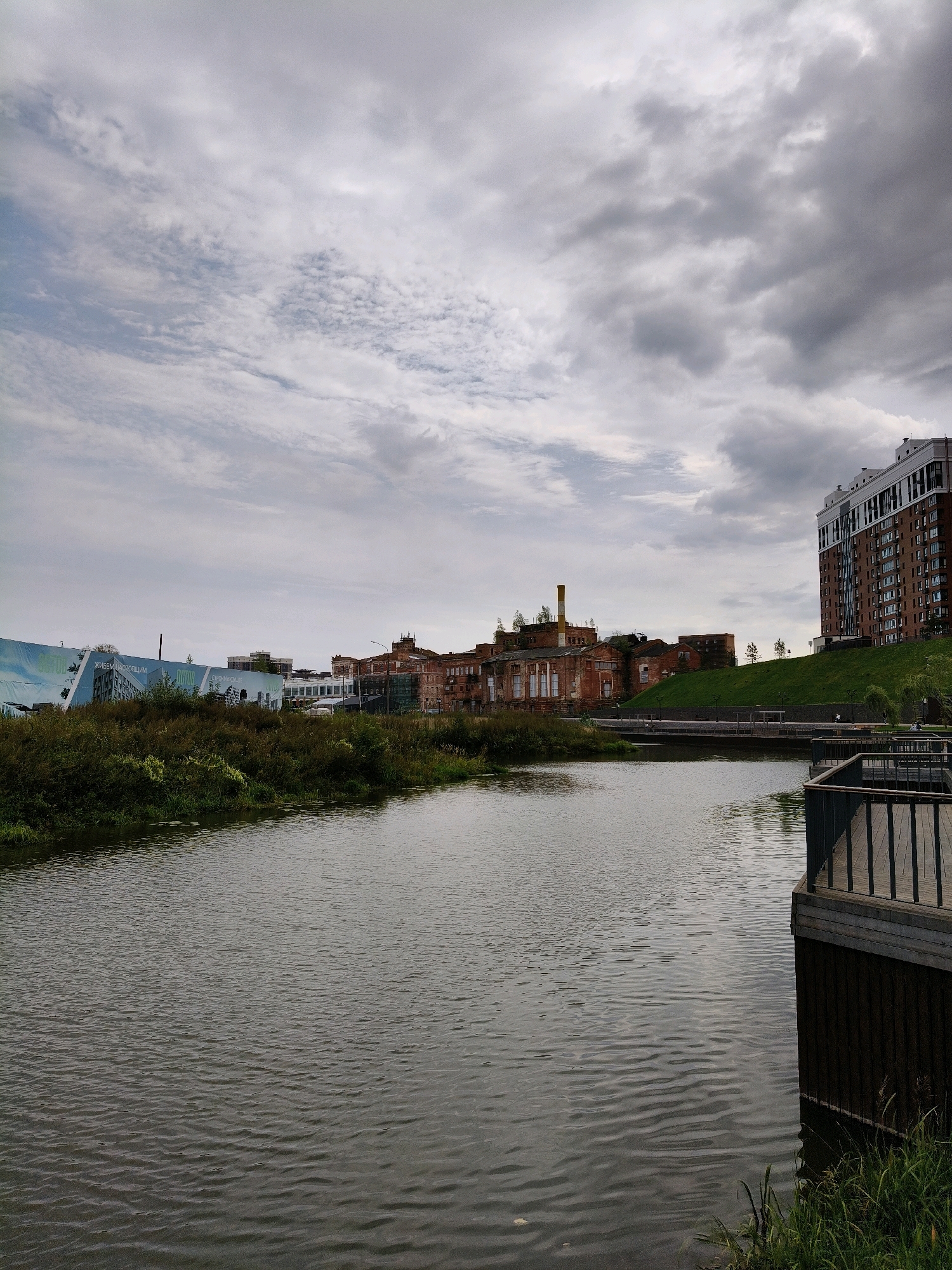 The image shows a wide, calm river or body of water in the foreground, with ripples on its surface. On the right side of the foreground, a wooden pier or deck juts out over the water, with a railing. Beyond the water, on the far bank, is a mix of old industrial buildings and a modern, multi-story residential building. The old buildings appear to be brick, with some showing signs of disrepair. A tall, yellow smokestack is visible on one of the older structures. To the left of the old industrial buildings, a large, modern glass structure is visible, partially obscured by vegetation. A grassy slope with some trees leads up to the modern residential building on the right. The sky is overcast with grey clouds. The scene appears to be a former industrial area that is being redeveloped or has been integrated into a park or recreational space. The location is identified as Naro-Fominsk, Russia.