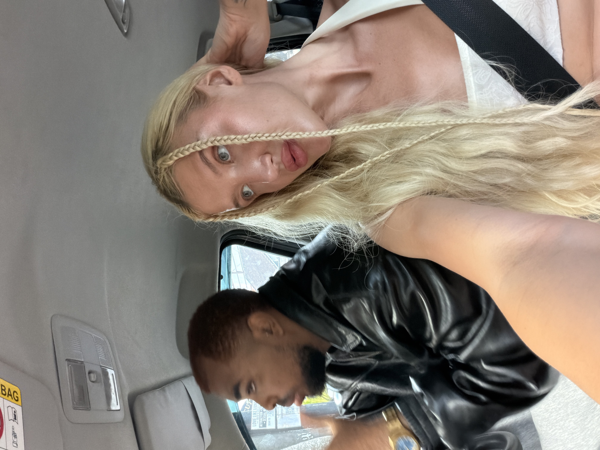 In a car in Mengwi, Indonesia, a person with brown skin and short brown hair, wearing a black leather jacket and a gold watch, drives. Beside them, a person with blonde hair styled in braids, wearing a white top, looks towards the camera. The car's interior is visible, including the roof lining and a seatbelt.