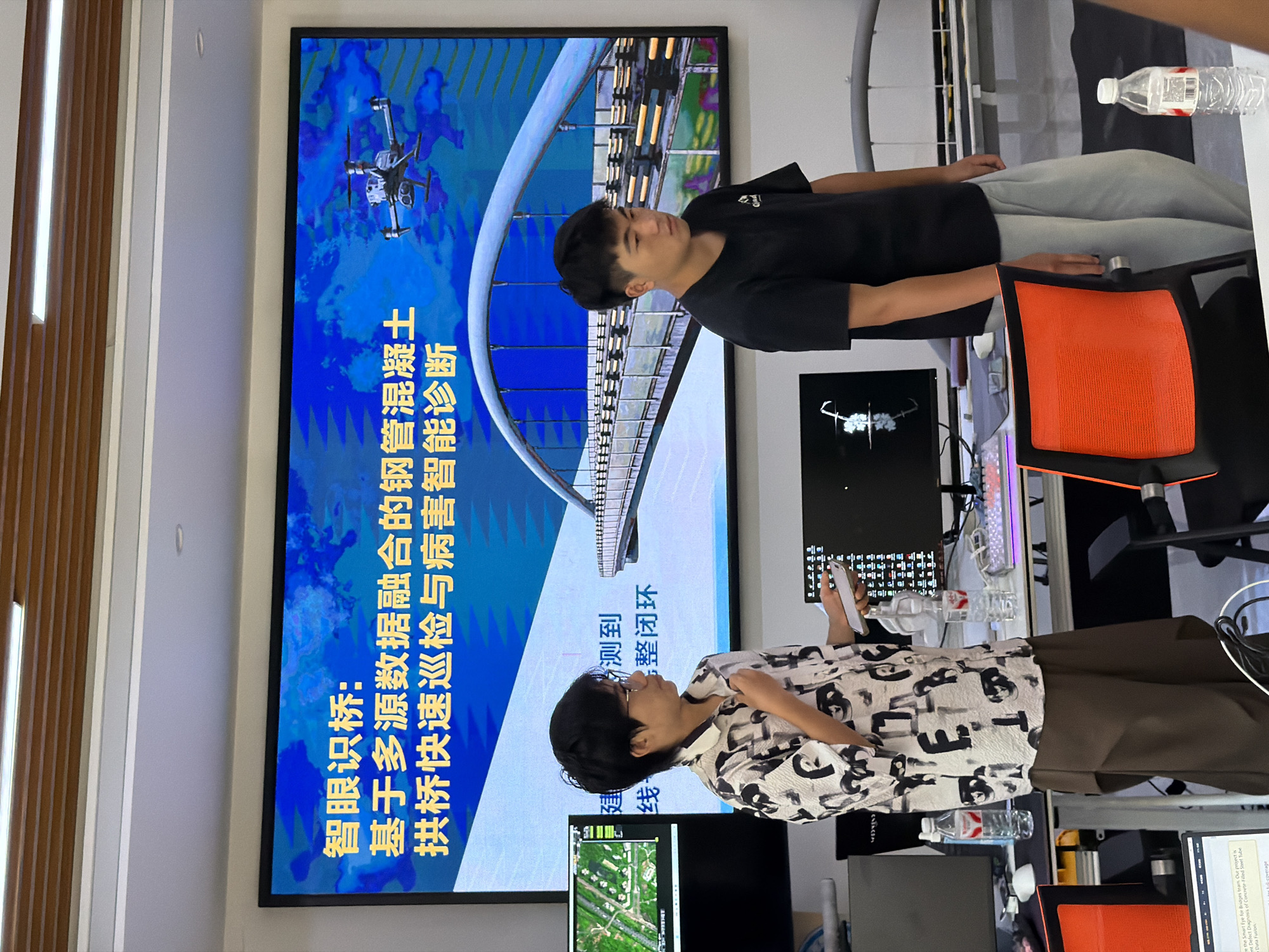 In an indoor setting, likely an office or presentation room, two young individuals stand before a large display screen. The screen exhibits a vibrant blue background with Chinese text, and an image of a bridge, a drone, and some abstract patterns. The text appears to be a title and description related to a project.
To the left, a person with short dark hair, glasses, and a patterned black and white shirt is gesturing with their right hand while holding a mobile phone. They are facing towards the screen, appearing to present or explain something.
To the right, a person with short dark hair, wearing a black t-shirt and light grey pants, is looking towards the left, seemingly listening to the other person.
In the foreground, desks are visible with computer monitors, keyboards, and bottles of water. The overall environment suggests a technical or research-oriented presentation. The location is identified as Jiangjin, China.