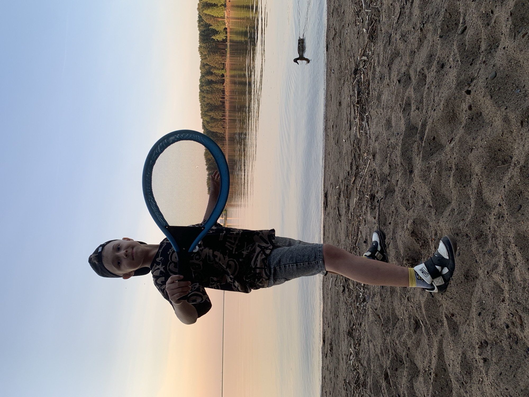 A young boy stands on a sandy beach holding a tennis racket. The water from a lake comes up to the beach. A treeline is visible across the lake. The sky is a light blue and pink. A single duck swims in the lake. The location is Chaikovsky, Russia.