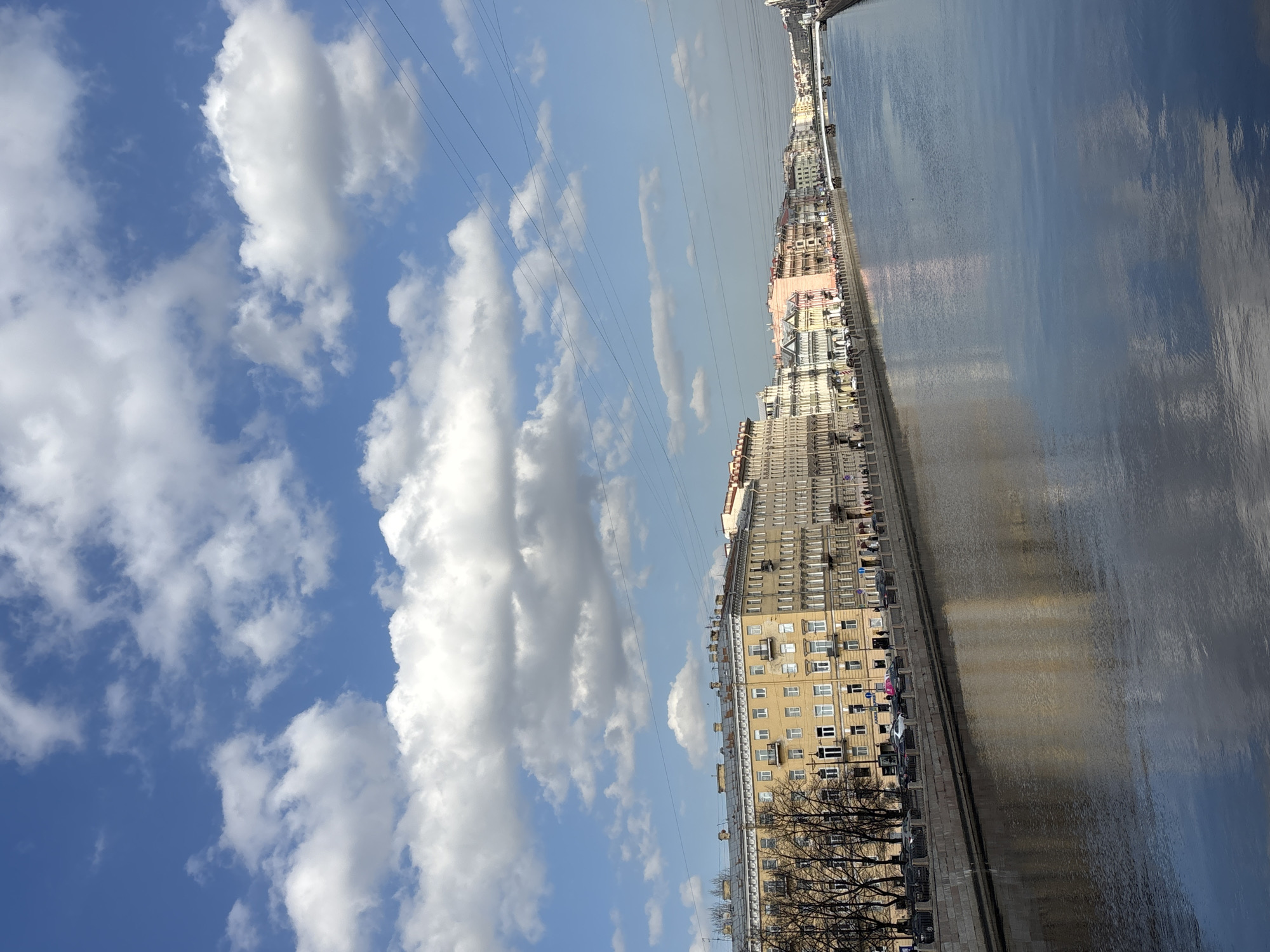 The image displays an urban waterway, specifically a canal or river, within the city of Saint Petersburg, Russia, under a clear blue sky with scattered white cumulus clouds. Bright daylight illuminates the scene, casting reflections on the water's surface.
The waterway, which occupies the lower half of the vertical frame, is relatively wide and features calm, dark blue water reflecting the sky and adjacent buildings.
Along the left bank, a stone embankment runs parallel to the waterway. Above the embankment, a row of bare deciduous trees with dark, slender branches is visible, suggesting late autumn, winter, or early spring. Beyond the trees, a sidewalk hosts several indistinct pedestrian figures walking in various directions. A street runs parallel to the sidewalk, populated by several parked vehicles, primarily dark-colored, with at least one white and one pink car identifiable.
Multi-story buildings line the street. The most prominent building in the mid-ground on the left is light yellow with numerous evenly spaced windows, many featuring white frames, and several small, dark balconies. Other buildings further down the embankment display varying pastel shades, including grey, light pink, and white, consistent with classical Saint Petersburg architecture.
In the distance, the waterway curves slightly, and at least two vehicular bridges span the water. The architecture of the distant buildings becomes less distinct but maintains a consistent style.
Several thin, dark power lines traverse the upper right quadrant of the sky, extending diagonally across the frame.