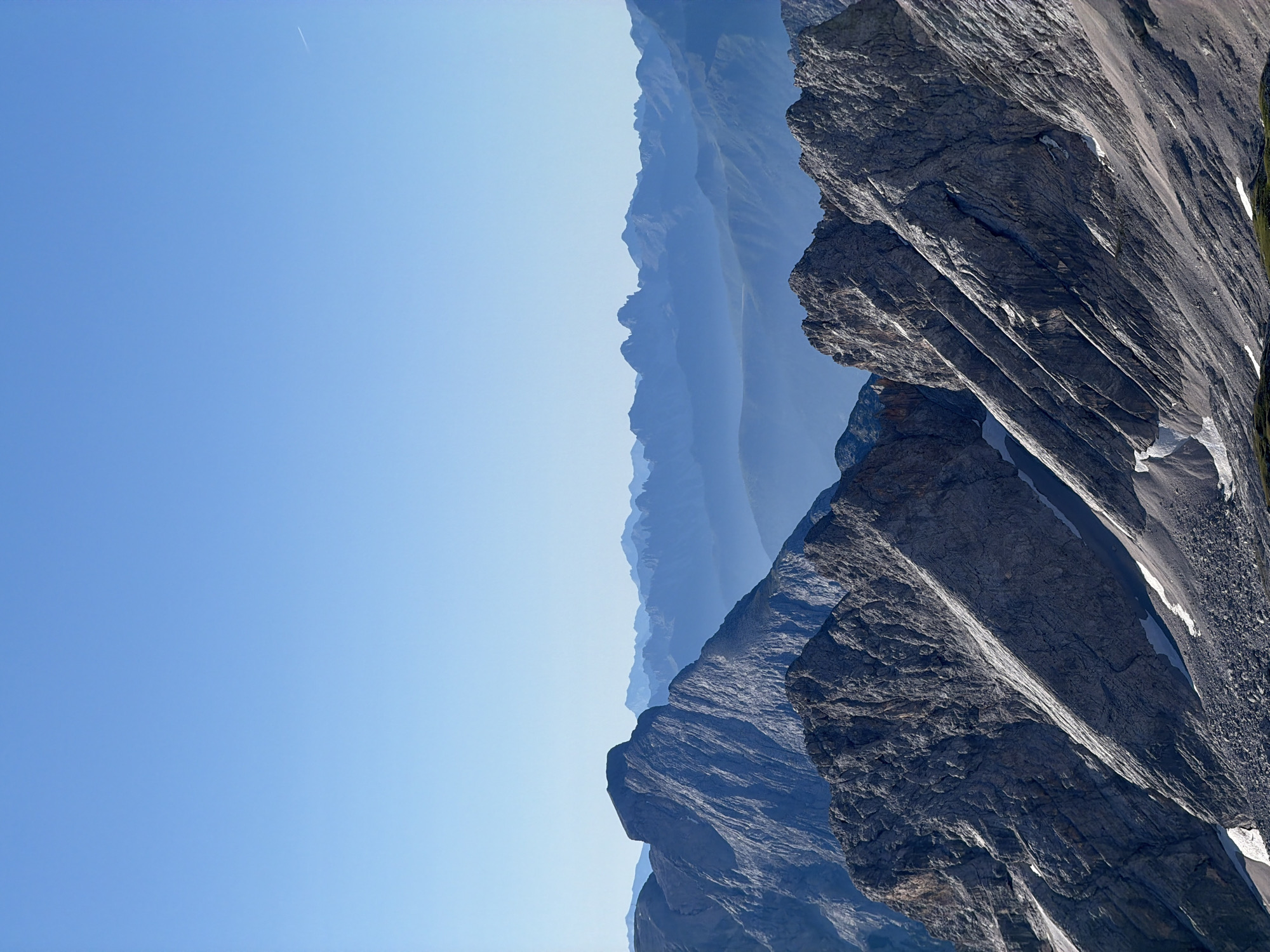 This is a high-angle, vertical, wide shot of a mountain range under a clear blue sky. The foreground features rugged, rocky mountain peaks with patches of snow. In the mid-ground, several layers of hazy, blue-toned mountains recede into the distance. The background is dominated by the vast expanse of the clear sky. This scene likely depicts the Alps, potentially near Garmisch-Partenkirchen, Germany, given the typical geological formations and atmospheric conditions of the region. No people or man-made objects are visible.