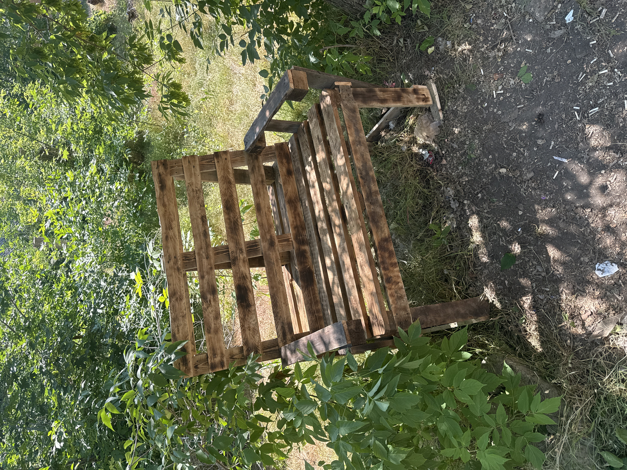 A rustic, wooden bench made from pallets sits outdoors in Makiivka, Ukraine. The bench, stained a dark brown, has a slatted seat and backrest, and simple armrests. It is positioned on a patch of dry grass and dirt, with a scattering of debris, including cigarette butts and wrappers, on the ground around it. Lush green foliage from surrounding trees and bushes partially frames the bench, casting dappled shadows on the scene. The lighting suggests it is daytime, with bright sunlight filtering through the leaves. There are no people visible in the image.