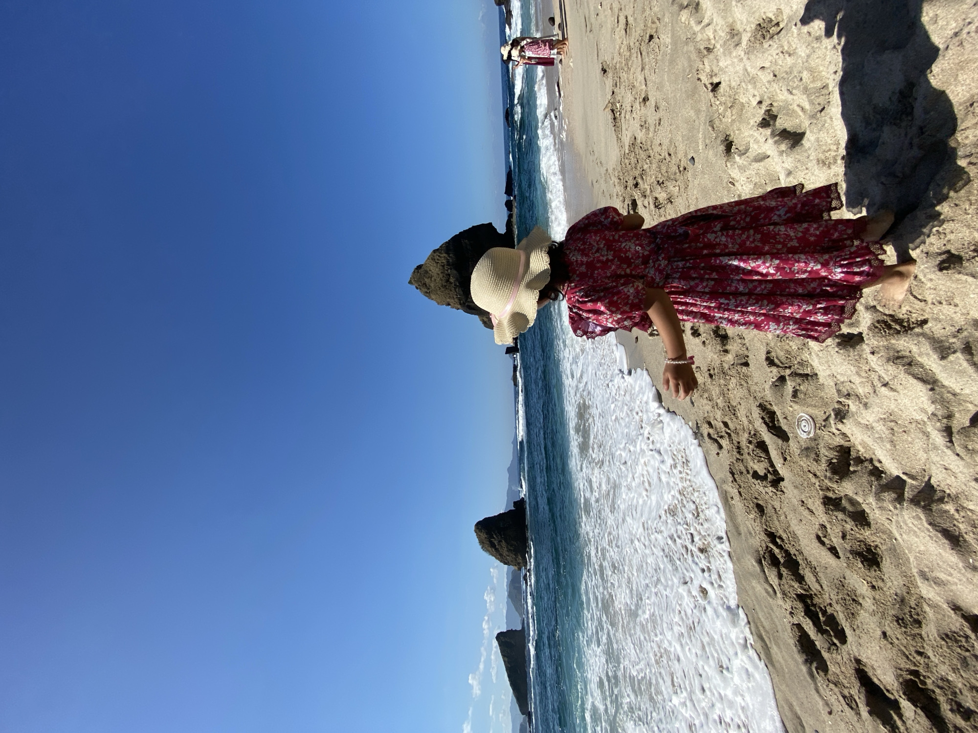 A medium shot depicts a beach scene on a clear day. A person wearing a red floral dress and a straw hat stands on the sandy beach. The person's bare feet are near the water's edge where waves are breaking. To the person's left, rock formations jut out from the ocean. In the distance, other people can be seen on the beach. The sky is a clear, bright blue. An object, likely a white plastic lid, can be seen in the sand. The location is Unknown, Indonesia.