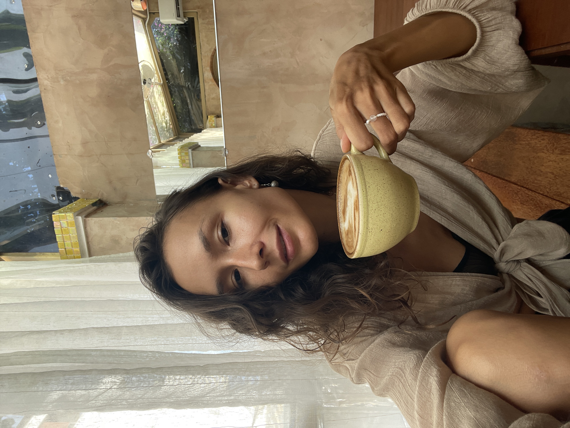A young woman with long, wavy dark hair smiles subtly at the camera while holding a light yellow-green ceramic mug containing a latte or cappuccino with visible latte art. She is wearing a light-colored, possibly textured linen or cotton top that appears tied at the front. A delicate beaded ring is visible on her right ring finger, and she has a small drop earring in her left ear.
The setting appears to be a brightly lit indoor space, possibly a cafe or a relaxed room. Natural light streams in from the left, partially obscured by sheer white curtains. Behind the woman, a light beige wall is visible. To the far left, there are reflections in a dark, glossy surface that seem to show an outdoor area with green foliage. Through a window or opening in the mid-background, part of an exterior structure with yellow mosaic tiles and more green vegetation can be seen. The architectural details and tropical foliage are consistent with the provided location context of Mũi Né, Vietnam. The time of day is clearly daytime, indicated by the abundant natural light. The woman's expression conveys a sense of calm and enjoyment. There is no visible text in the image.