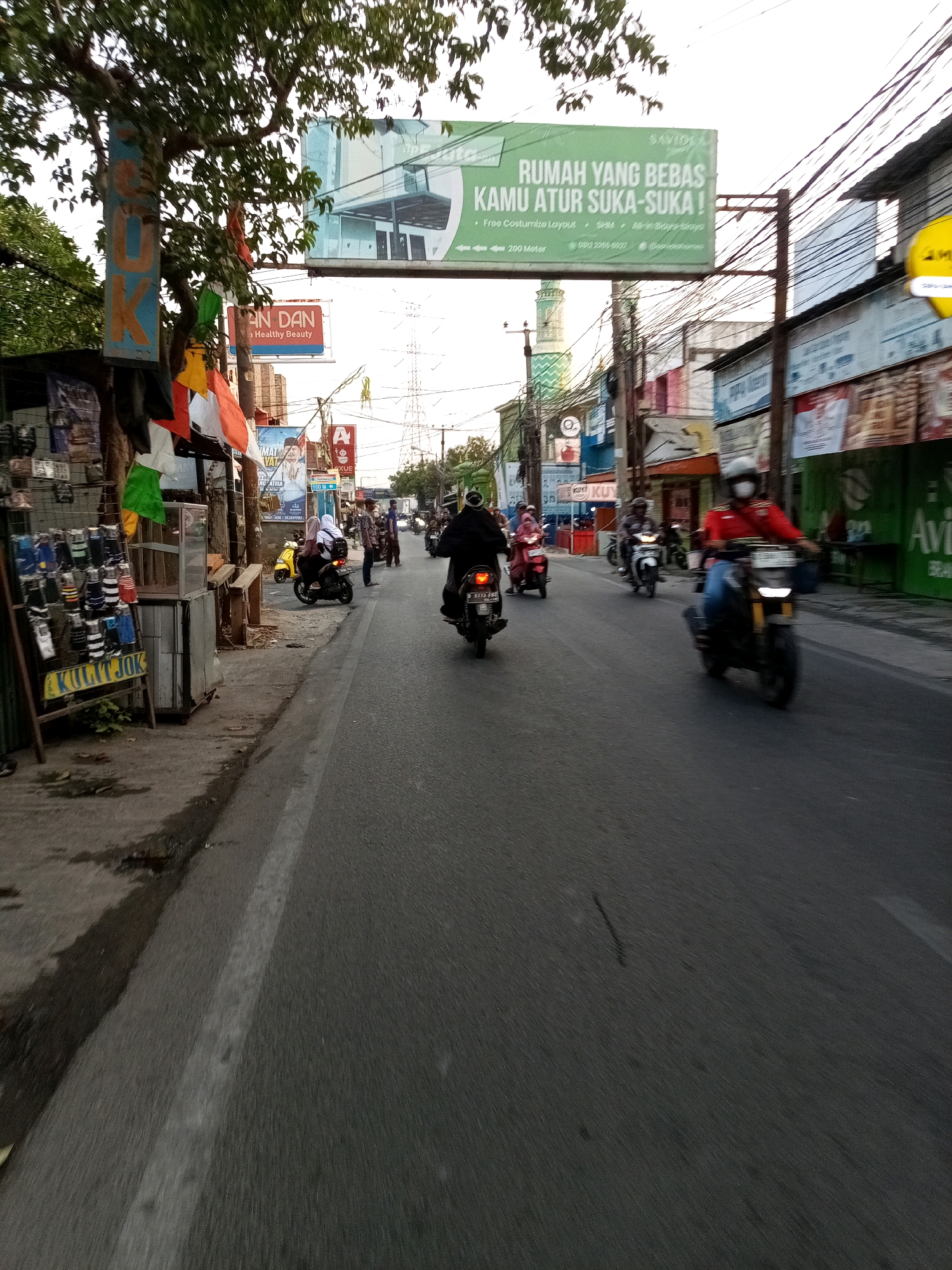 The image depicts a street scene in Karangsatria, Indonesia, with a focus on a road with several motorcycles and riders. The road is asphalt, with a white line marking the lane. On the left side of the road, there is a sidewalk with various objects, including a rack of what appear to be motorcycle seat covers labeled "KULIT JOK," and some flags or banners hanging from a tree. Buildings line the right side of the street, featuring storefronts and signage, including a large green billboard with text in Indonesian that reads "RUMAH YANG BEBAS KAMU ATUR SUKA-SUKA !" which translates to "A house you can freely arrange as you like!". A green minaret of a mosque is visible in the background. Several motorcycles are in motion, some with riders wearing helmets and casual clothing. One motorcycle in the foreground is red and black, and another rider is wearing a red jacket and a white face mask. The overall atmosphere suggests a typical urban environment during daylight hours.