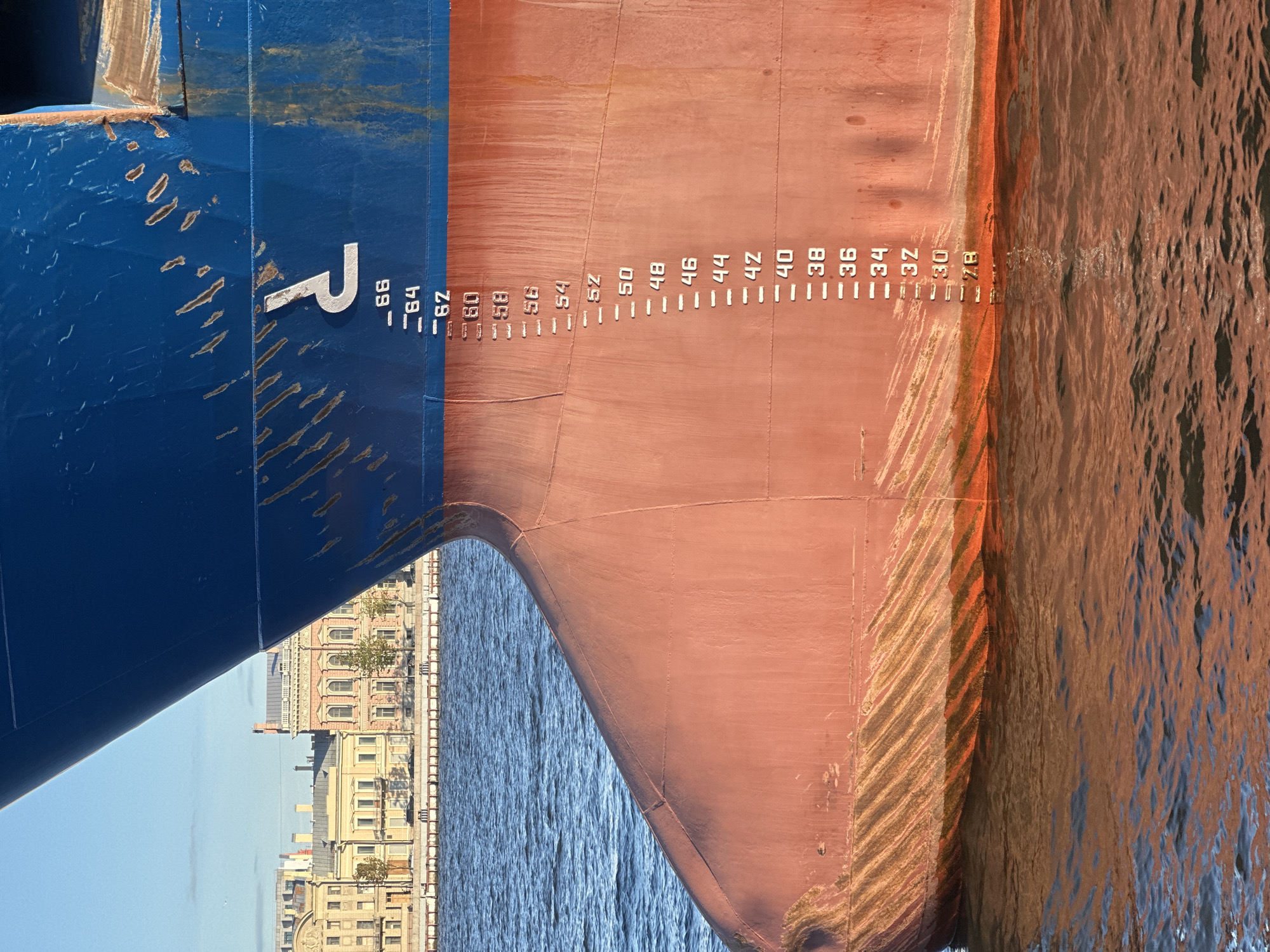 A close-up, medium shot captures the bow of a large blue and red ship docked in the Neva River in Saint Petersburg, Russia. The ship's hull is marked with white numbers, indicating draft measurements, with the water level reaching the mark labeled "32". In the background, a row of classic, colorful buildings with mansard roofs line the riverbank. The sky is a clear, pale blue. The image focuses on the details of the ship's hull and the architectural style of the buildings.
