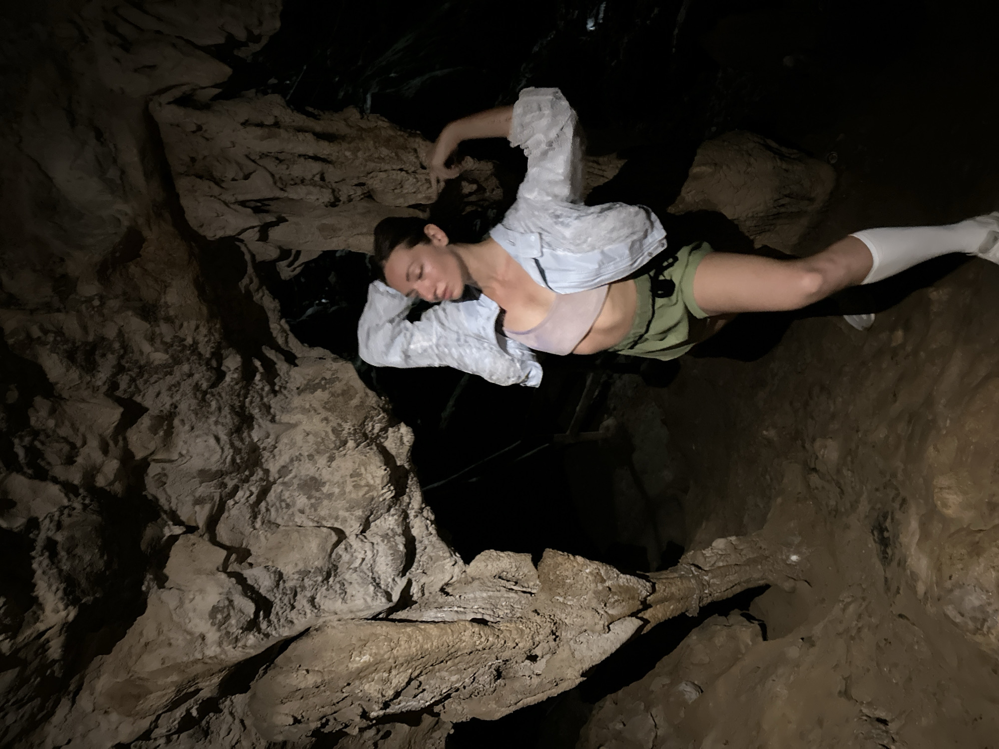 A young woman with dark hair pulled back stands in a dimly lit cave, her eyes closed and head tilted back as she runs a hand through her hair. She wears a light blue, textured button-down shirt over a pink bralette, green shorts, white knee-high socks, and white sneakers. The cave walls are rough and textured, with stalactites hanging from the ceiling and stalagmites rising from the ground, characteristic of the limestone karst landscape of Mai Chau, Vietnam. The lighting suggests artificial illumination, likely from a flashlight or portable lamp, casting shadows and highlighting the geological formations.