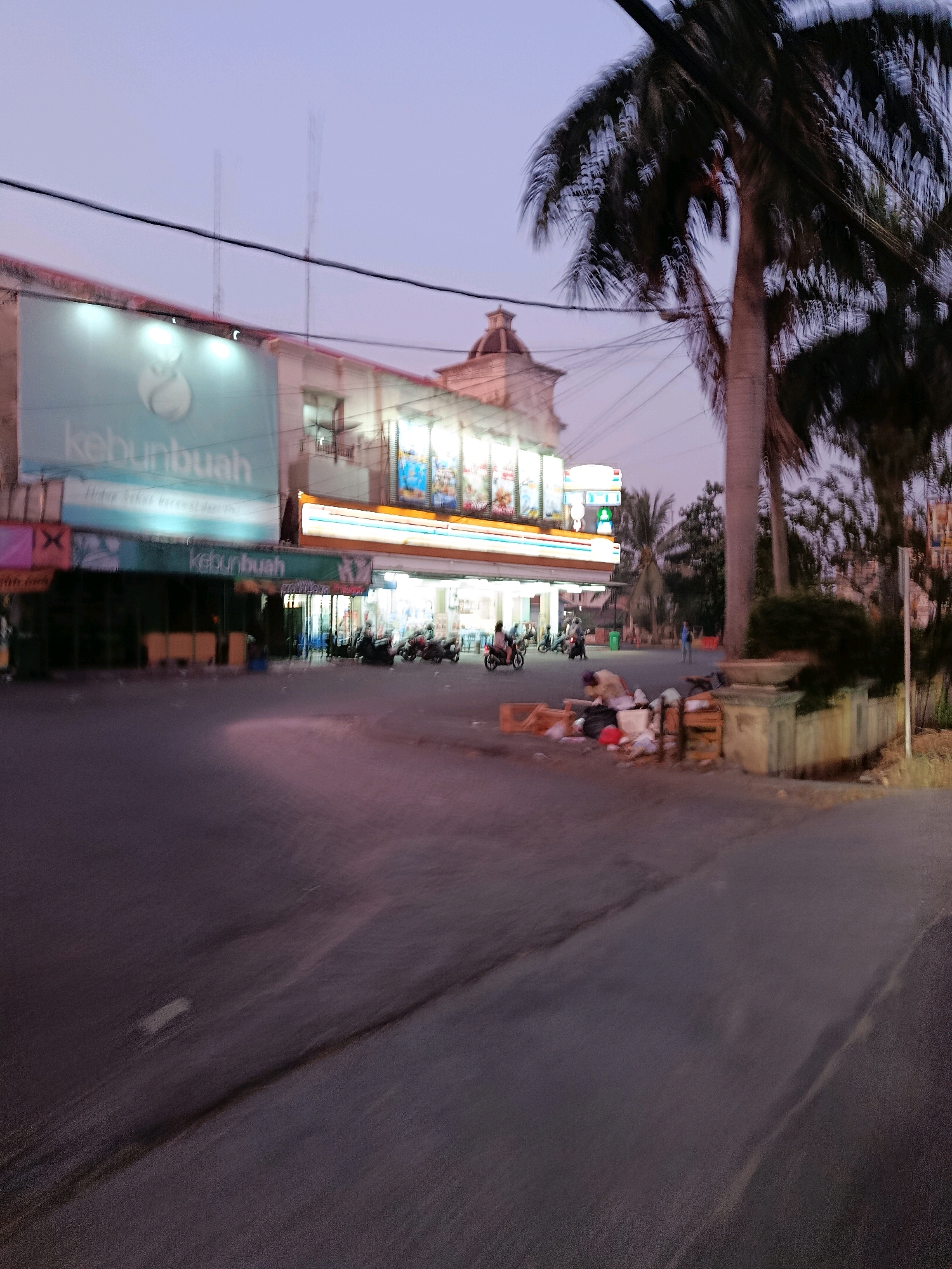 The image captures a street scene in front of a commercial building at dusk, likely in Bekasi, Indonesia. The building features a large illuminated sign with the text "kebunbuah." Adjacent to it, a 7-Eleven convenience store is visible, with its bright lights illuminating the storefront. Several motorcycles are parked in front of the stores, and a few individuals are present, though their actions are not clearly discernible due to motion blur. On the right side of the image, a pile of trash and wooden crates is scattered on the ground next to a low wall. Palm trees are prominent in the background, characteristic of the tropical climate. The sky is a soft, hazy purple, indicating twilight. The street itself is paved and appears to be a typical urban thoroughfare. The overall impression is one of a bustling commercial area transitioning into the evening.