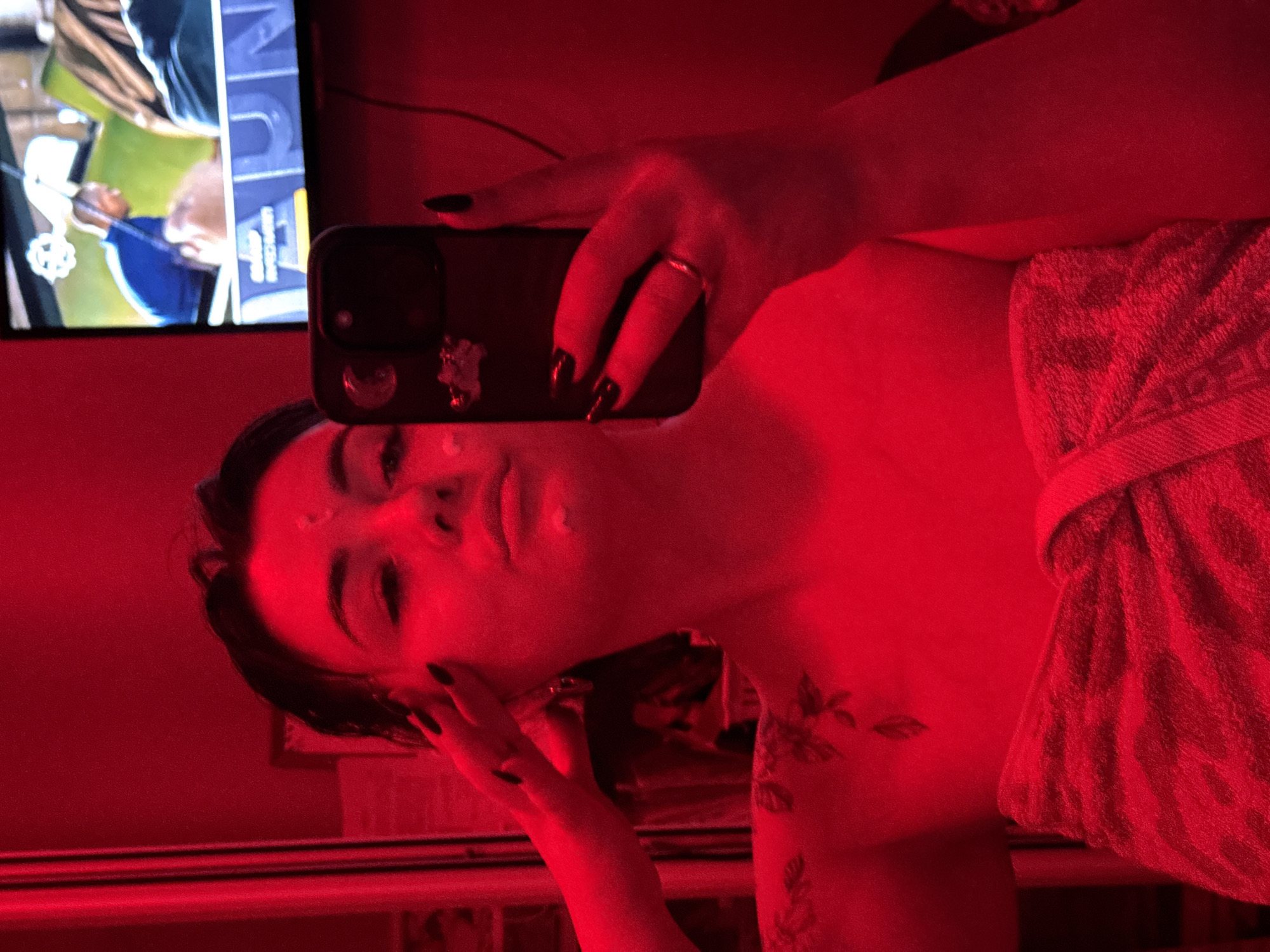 A woman is taking a selfie in a room with red lighting. She has on a towel and appears to have lotion or cream on her face. A TV screen is visible in the background displaying a soccer game. The location is inferred from metadata to be the city of Chaikovsky, Russia, though no specific features in the image confirm this.