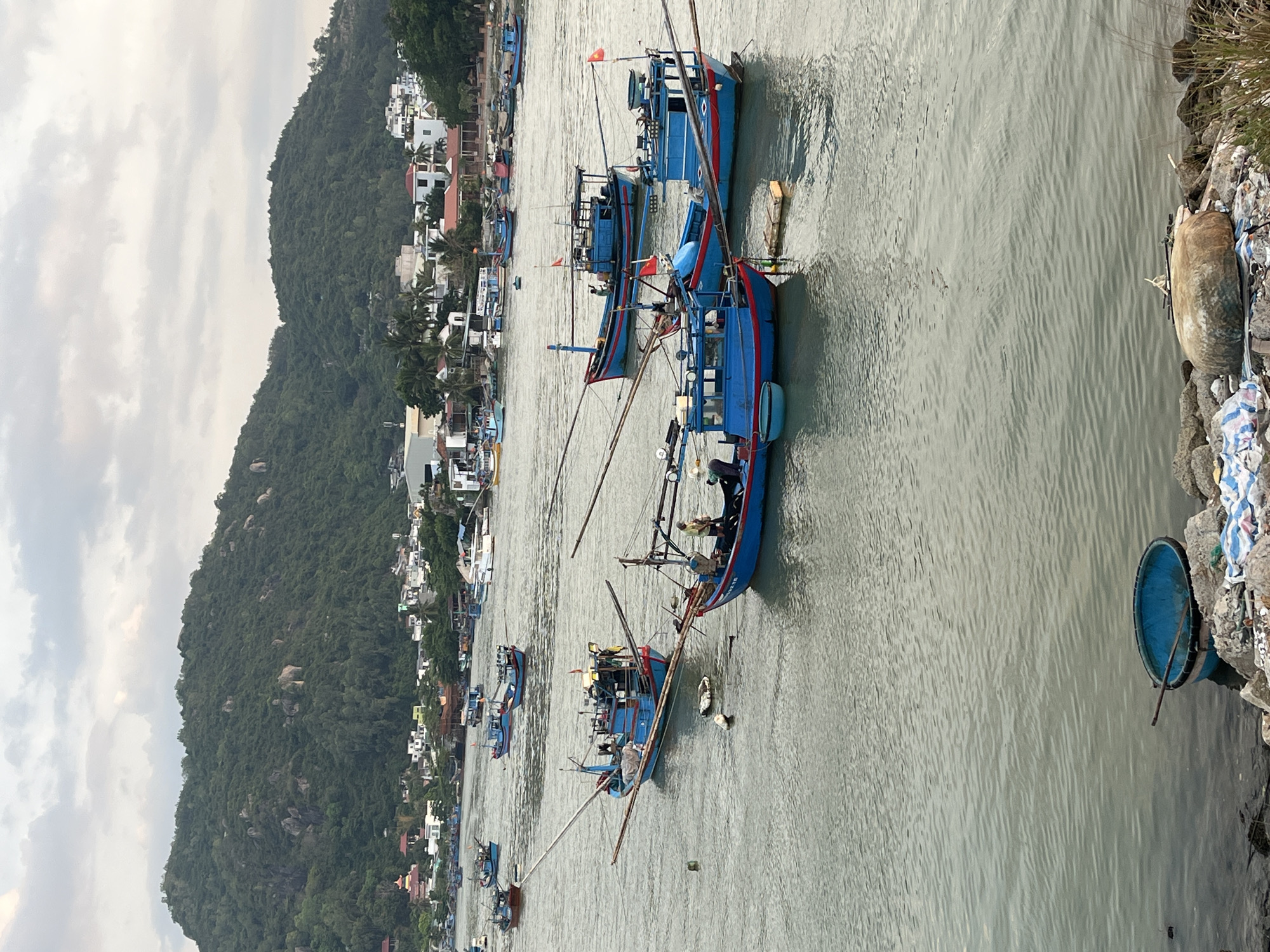 The scene depicts a body of water, likely a river or bay, adjacent to a coastline with a dense green hill ascending in the background. On the left, a town is nestled between the water and the hill, featuring buildings of varying sizes and colors. In the water, several blue fishing boats with long, thin masts are visible. Some of the boats have occupants who appear to be engaged in activities onboard. The sky is overcast with cloud cover. A shoreline is located on the right side of the image and contains various refuse. The location is likely Bac Nha Trang Ward, Vietnam, given the geography and the type of boats present.