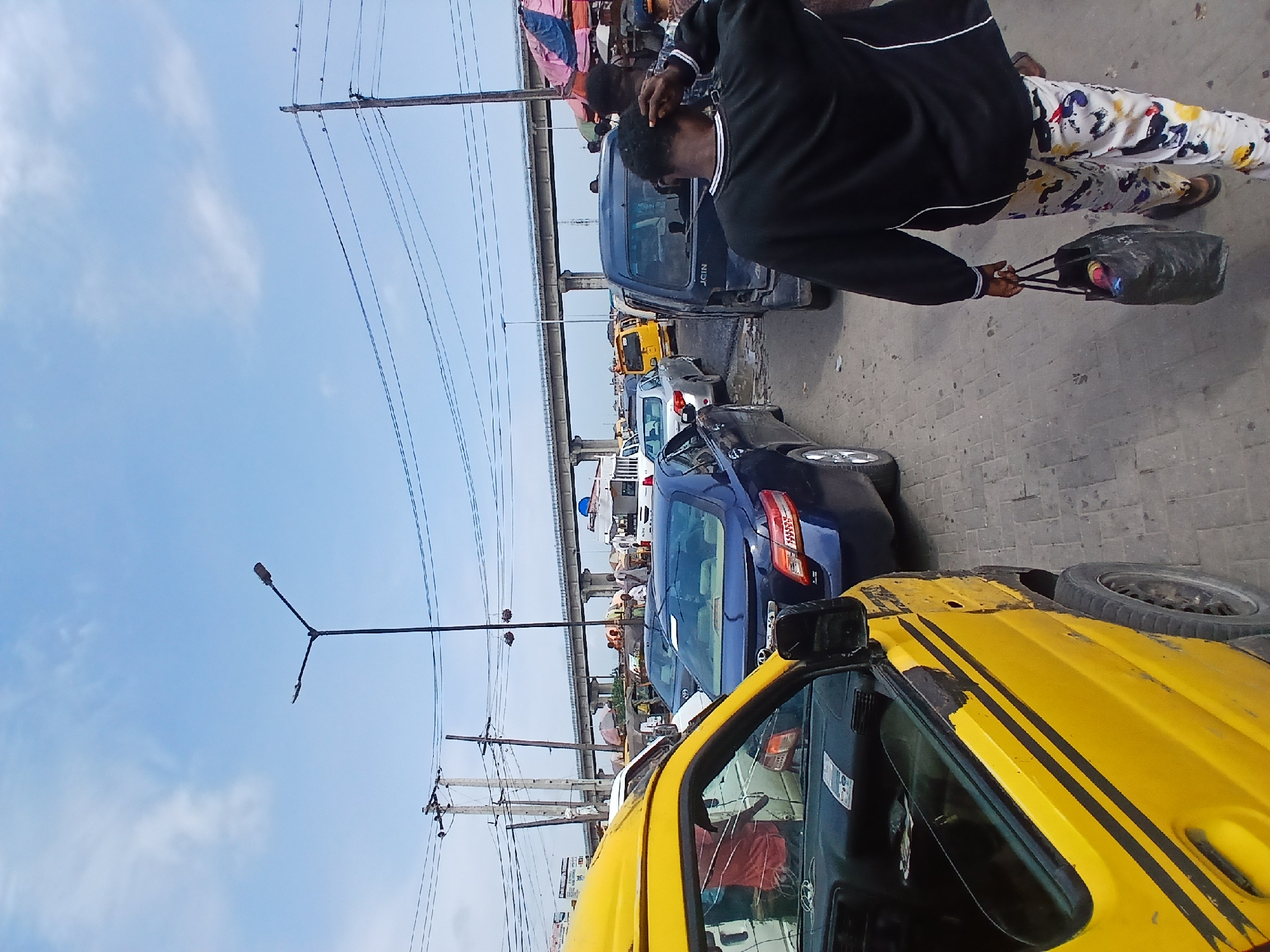 The image depicts a busy street scene, likely in Agano, Nigeria, characterized by heavy traffic and urban infrastructure. In the foreground, a yellow commercial vehicle is partially visible on the left, showing its side door and tire. Moving towards the background, a procession of cars and vans fills the roadway, indicating a traffic jam. Among the vehicles, a dark blue sedan is prominent, followed by a dark colored van with the word "JOIN" on its back. A person in a black shirt and brightly patterned pants is walking away from the camera, carrying a black plastic bag. Further down the street, more vehicles and pedestrians are present, with market stalls visible under a large elevated highway or bridge structure that spans across the scene. Power lines and utility poles are also visible, contributing to the urban setting. The sky is partly cloudy, suggesting daytime.
