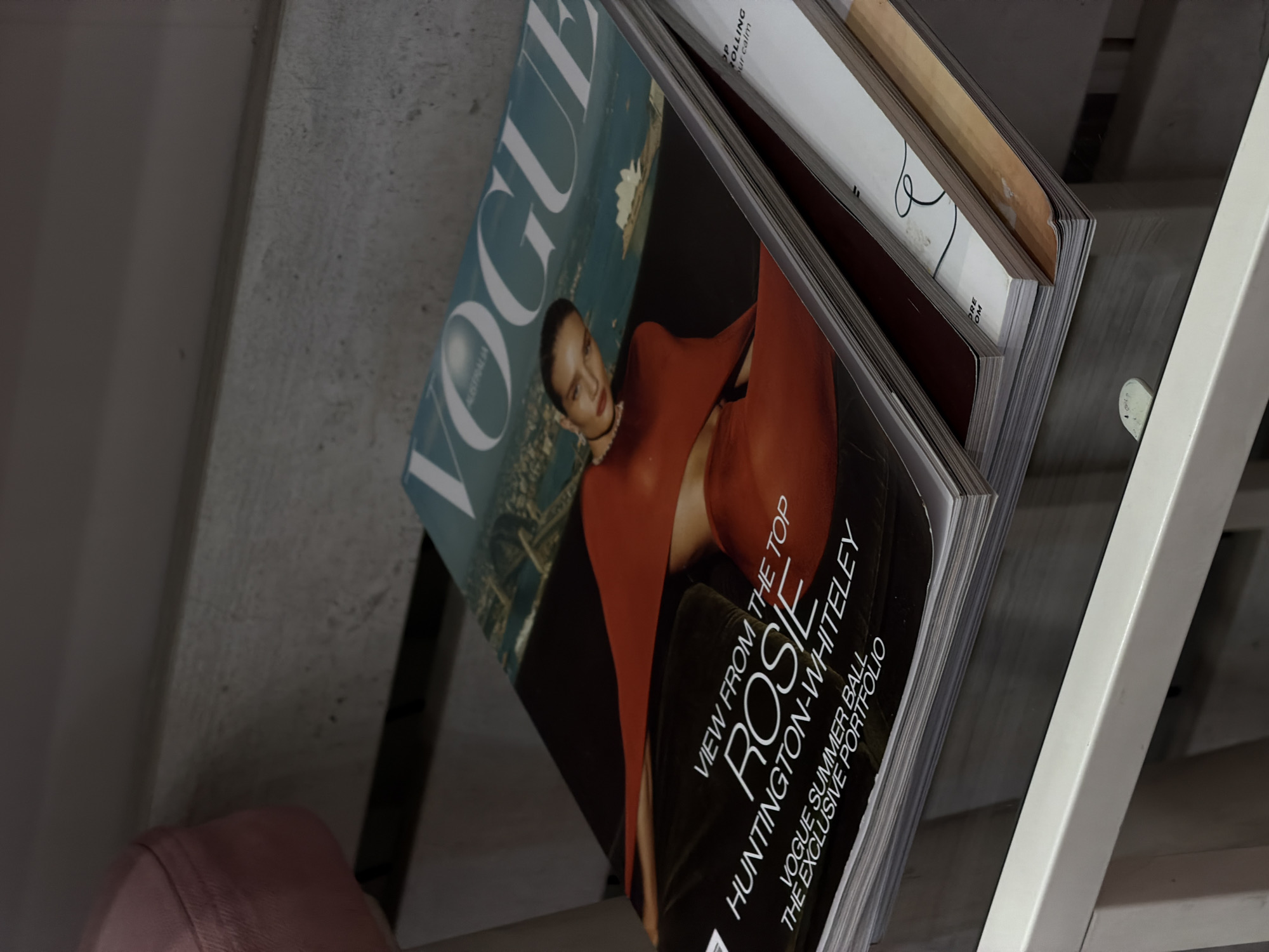 A stack of fashion magazines, including a copy of Vogue Australia featuring Rosie Huntington-Whiteley on the cover, rests on a glass table. The cover image shows Huntington-Whiteley in an orange outfit with the Sydney Opera House visible in the background. Other magazines are stacked beneath the Vogue issue. The scene appears to be indoors, possibly in a home or office setting, with a hint of a pink upholstered chair to the left. The lighting is soft and diffused. There is no visible information indicating the location is in Bishkek City, Kyrgyzstan.