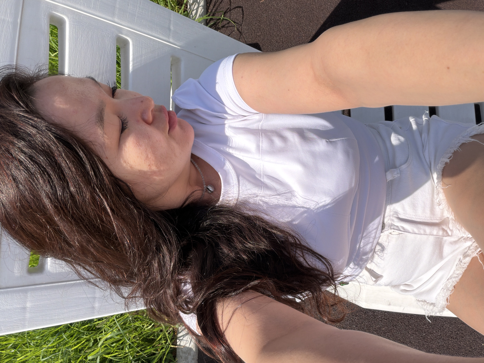 A woman with dark brown hair lies on a white bench outdoors. She wears a white T-shirt and white denim shorts. Her head rests on the bench, and her eyes are closed. The bench is positioned on grass, with a paved surface partially visible nearby. The scene is brightly lit, suggesting it is daytime. Location is unknown.