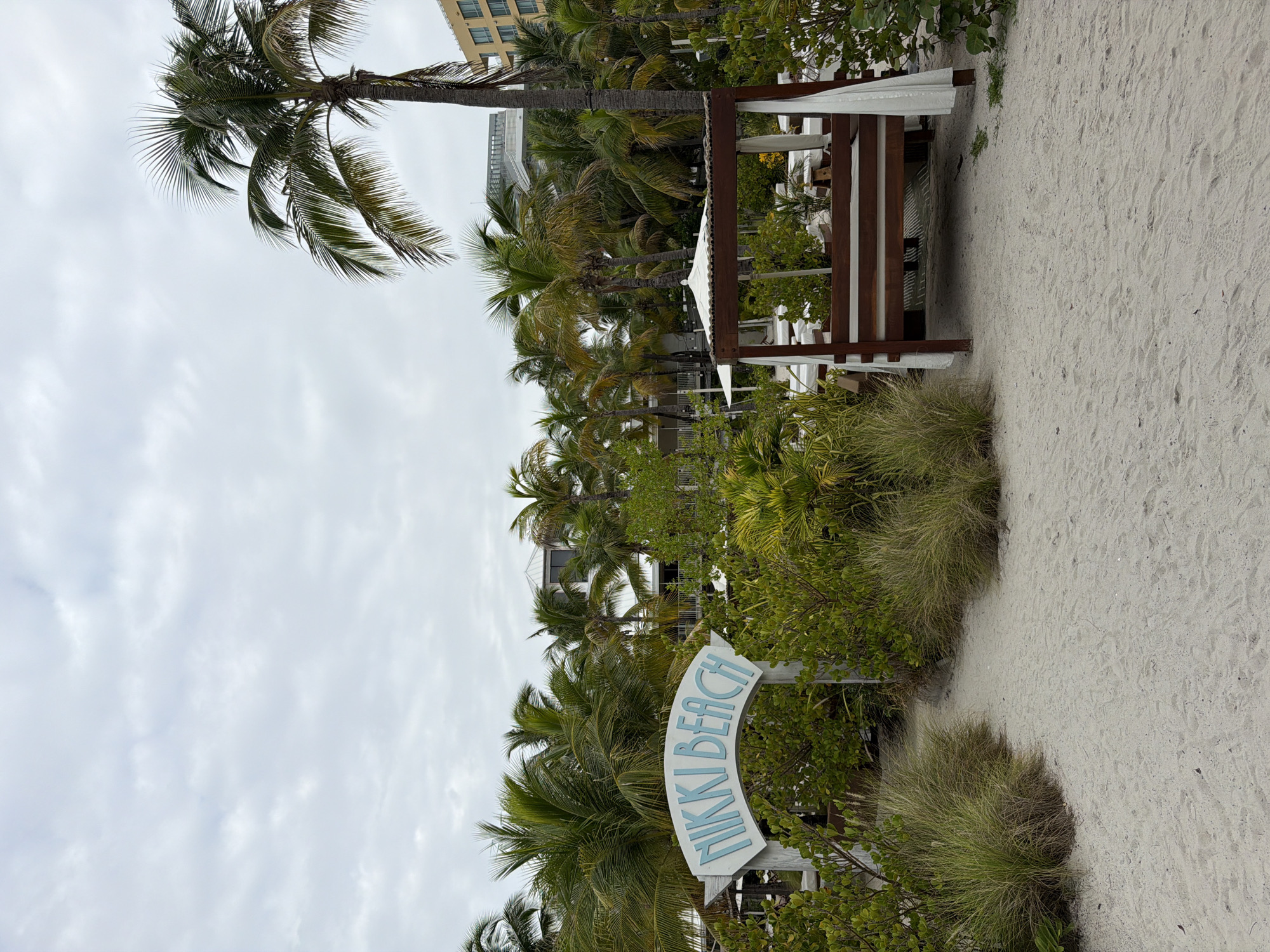 A beach pathway leads through lush green vegetation towards an archway with "NIKKI BEACH" written in light blue lettering. To the right, a wooden cabana with white curtains stands on the sandy beach. Palm trees are scattered throughout the scene, and buildings are visible in the background. The sky is overcast.