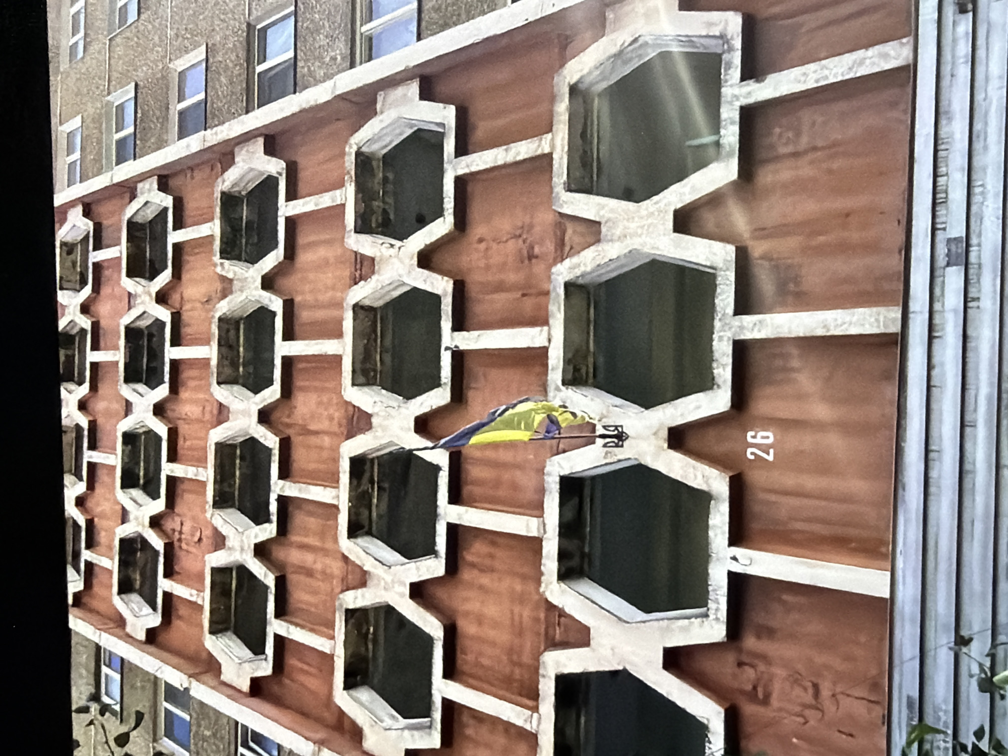 A vertical photograph captures the facade of a multi-story residential building, consistent with Soviet-era architecture, located in Kyiv, Ukraine. The main section of the facade displays a reddish-brown textured wall adorned with a grid of repetitive, light-colored, interlocking concrete elements forming deep, recessed openings, possibly balconies or loggias. To the right, a plainer concrete section of the building features multiple rectangular windows with light-colored frames. A Ukrainian flag, displaying blue over yellow, is affixed to one of the central facade elements, directly above a dark Ukrainian national emblem (Tryzub). The white numeral "26" is visible on the lower portion of the reddish-brown wall. The building exhibits signs of weathering and age, with variations in the facade's color and texture. No individuals are visible in the scene.