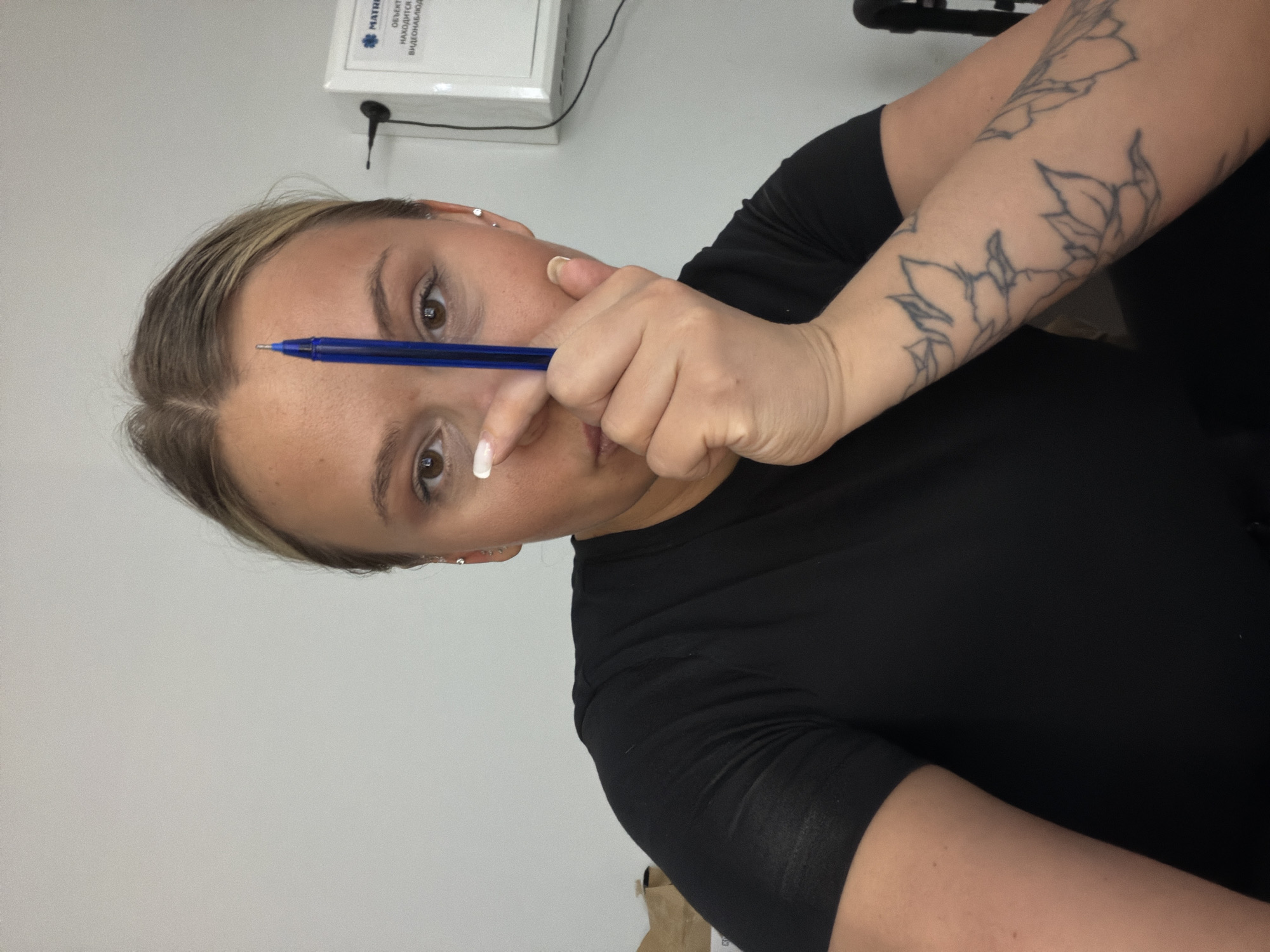 A woman with fair skin and light brown hair is holding a blue pen horizontally across her forehead. She is wearing a black shirt and has a floral tattoo on her left arm. She is looking directly at the camera. In the background, there is a white wall with a white electrical box mounted on it. The box has Russian text on it. The location is presumed to be in Sarapul, Russia, based on the presence of the Russian text.