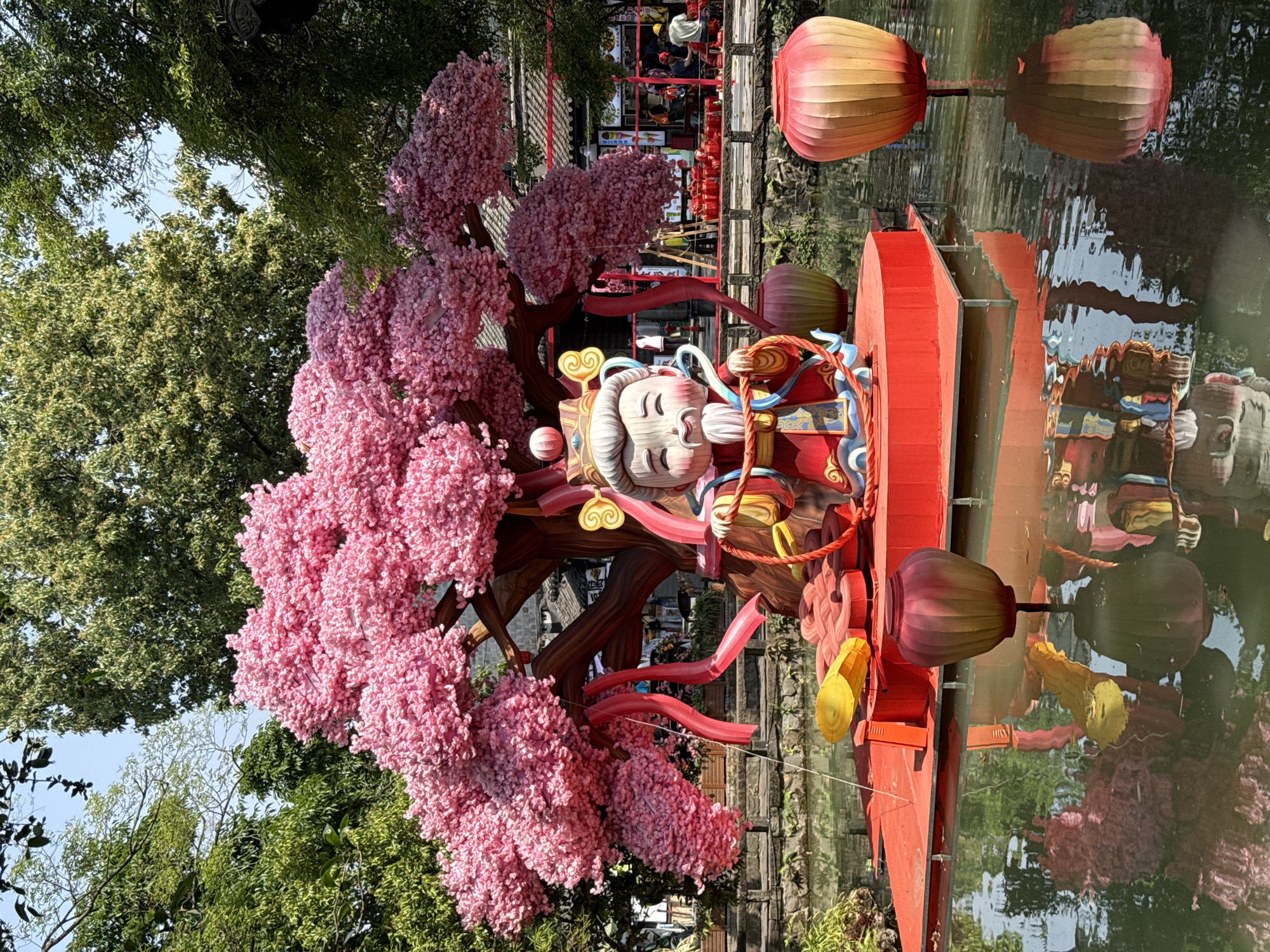 The image shows a colorful artistic display in an outdoor setting. A vibrant pink cherry blossom tree is in full bloom beside a red-painted structure holding a statue of an elderly woman with closed eyes and a serene expression. The woman is holding a rope-like object, possibly part of a decorative or ceremonial element. Lanterns in shades of orange and pink hang near the structure and reflect in the body of water. Buildings can be seen in the background. Given the elements visible in the image, the location is likely in Wuhou District, China.