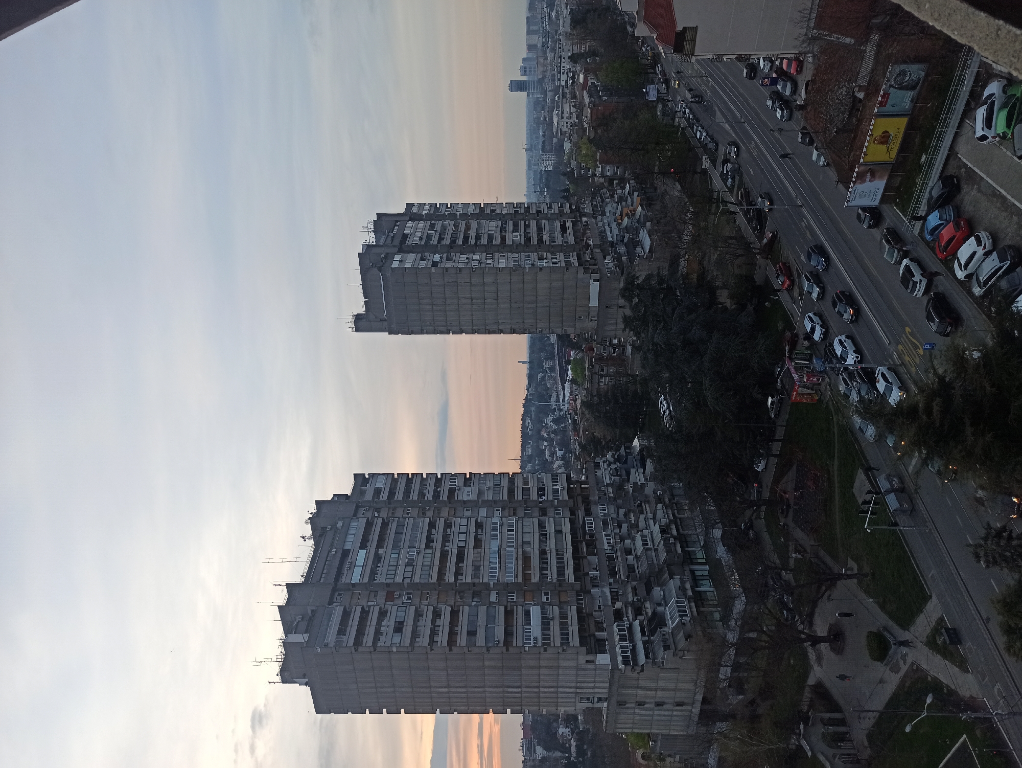 The image is an aerial, slightly rotated view of two large, concrete residential buildings in the Vozdovac Urban Municipality of Belgrade, Serbia. The buildings are similar in design, with a brutalist architectural style, featuring staggered levels and projecting balconies. Between the buildings is a view of the city and sky. To the right of the buildings is a busy street with cars parked along the sides and moving along the traffic lanes. A park and smaller buildings are visible closer to the camera. The sky is a mix of pale blue and orange. The overall lighting suggests it is dusk or dawn.