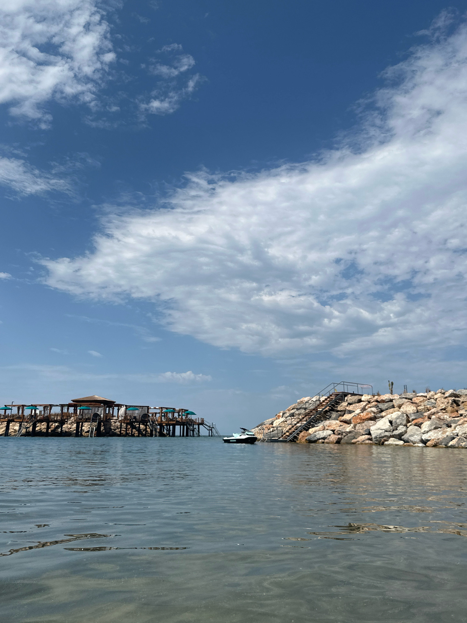 The image depicts a tranquil scene at the coast of Agios Epiktitos, Northern Cyprus. In the foreground, the clear, shallow water reveals the sandy seabed, with subtle ripples indicating a gentle breeze. The water is a translucent blue-grey, reflecting the sky above.
Dominating the left side of the mid-ground is a wooden structure built on stilts over the water. This structure features multiple levels, with thatched roofs providing shade over seating areas. Several turquoise umbrellas are visible, suggesting a place for relaxation or dining. Stairs lead down from the structure to the water.
To the right of the wooden structure, a man-made rocky embankment rises from the sea. A metal staircase with handrails ascends this embankment, leading to its crest. A jet ski is parked near the base of the rocky embankment, partially submerged in the water.
The background is characterized by a bright blue sky dotted with fluffy white clouds, some of which are wispy and stretched, while others are more dense. The overall impression is one of a serene coastal resort or a peaceful day at the beach. There are no people visible in the image.