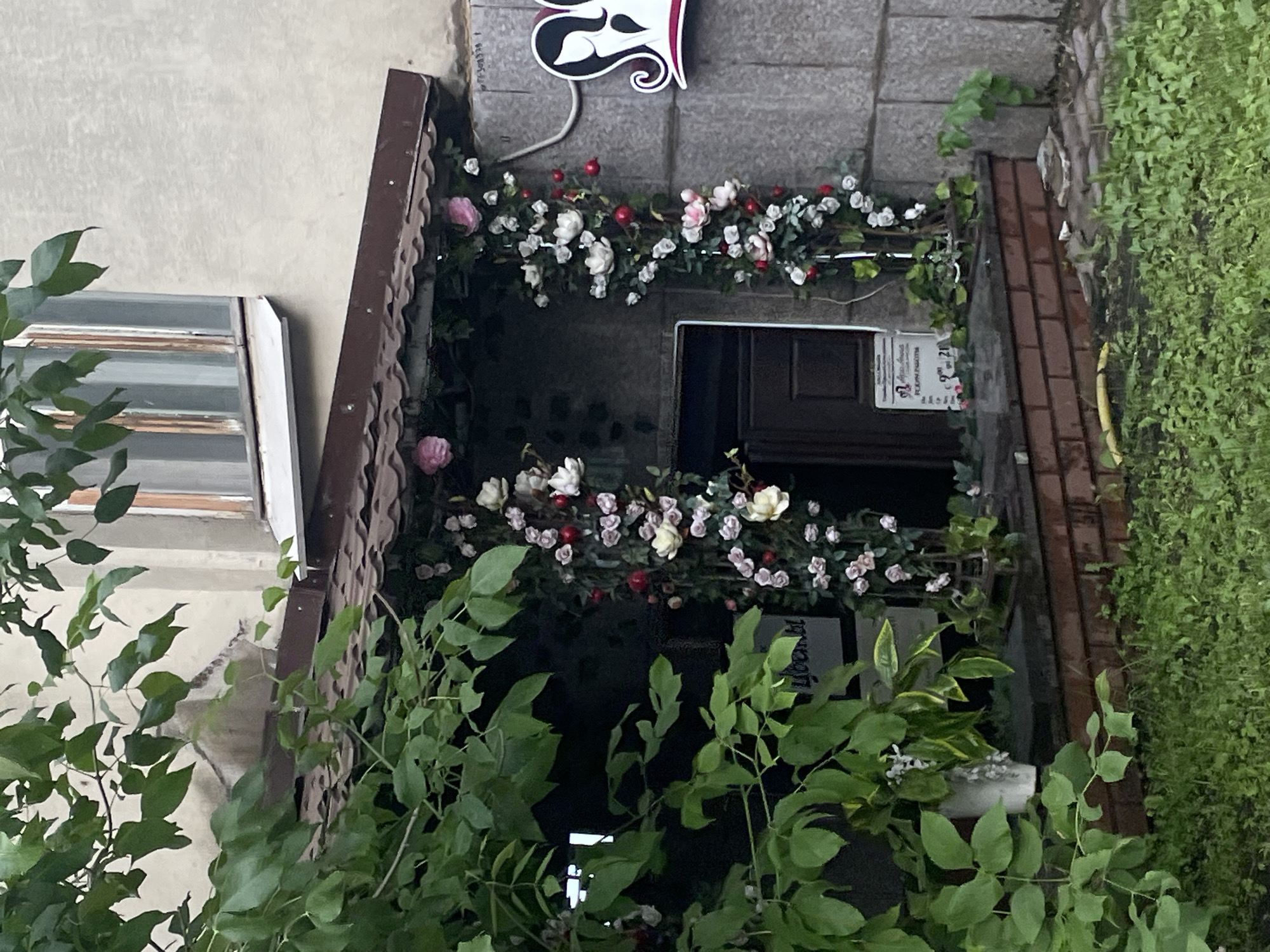 The image shows a storefront decorated with artificial flowers. The storefront is located on a street in Moscow, Russia. The building is made of concrete blocks, and the storefront has a brown tiled roof. The entrance is dark brown, and there are two pillars decorated with artificial flowers on either side of the entrance. A sign with text is visible on the left pillar, and another sign is on the ground to the right of the entrance. The storefront is surrounded by greenery, with large leafy plants in the foreground and grass growing on the ground. A logo with two female silhouettes is visible on the right side of the building. The overall impression is that of a flower shop or a similar business.