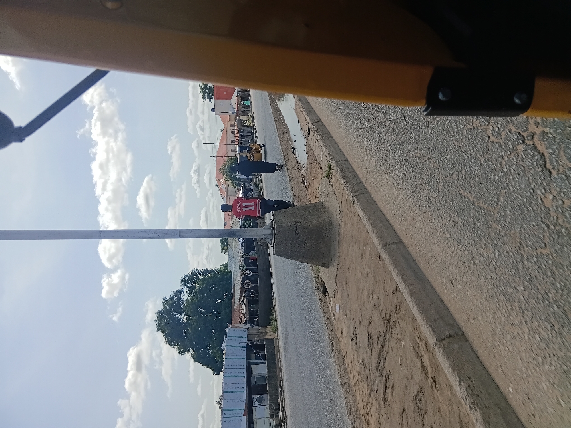 The image captures a street scene in Jalingo, Nigeria. In the foreground, a yellow vehicle is partially visible on the right, obscuring a significant portion of the frame. The road surface, which appears to be asphalt with visible aggregate, takes up the lower half of the image. A concrete curb separates the road from a sandy embankment on the left, where scattered debris is present.
Further down the road, two individuals are walking away from the viewer. The person on the left wears a red sports jersey with the name "ZEGRAS" and the number "11" on the back. The second individual, to the right, is dressed in dark clothing. A small, yellow motorized tricycle, commonly known as an "okada" or "keke," is visible further down the road in the middle lane, traveling in the same direction as the pedestrians.
On the left side of the road, a row of shops or market stalls are visible. These structures have corrugated metal roofs and are constructed with various materials, including what appears to be wooden planks and possibly some white sheeting on the front of one building. Objects resembling washing machines are displayed outside one of the shops. In the background, residential buildings with red-tiled roofs and trees can be seen under a partly cloudy sky. A tall, slender lamppost stands prominently in the middle ground, near the curb.
