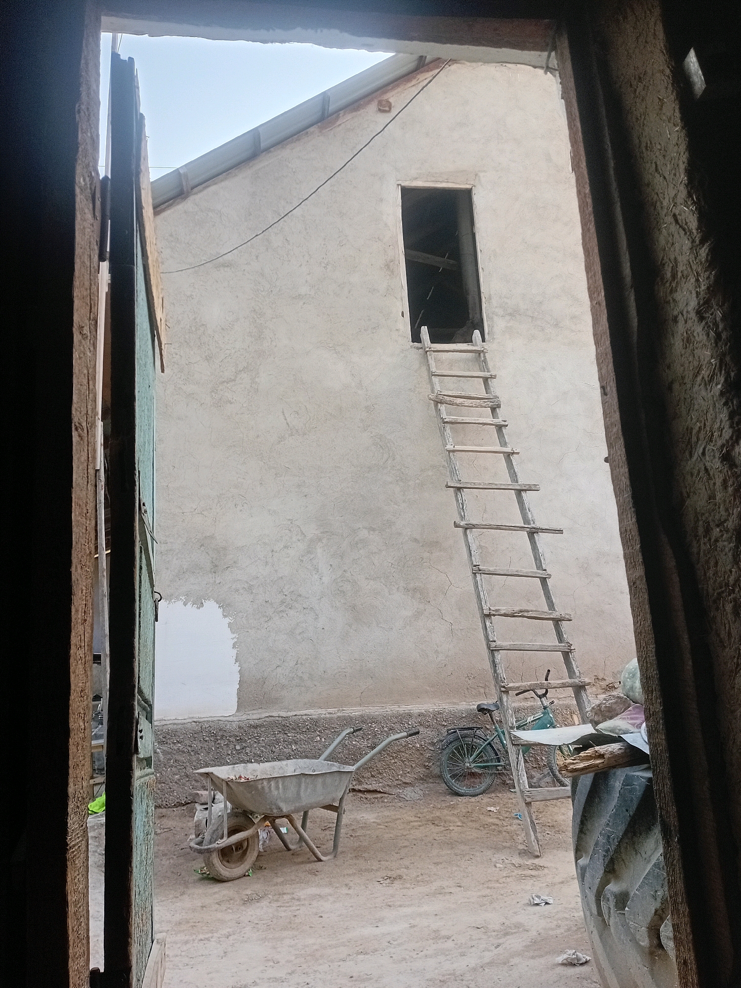 A wooden ladder leans against a light-colored, rough-textured building in Ажек, Kyrgyzstan. The ladder extends to an open doorway above. In front of the building, a green bicycle is parked. To the left of the bicycle, a wheelbarrow rests on the dirt ground, with debris in its basin. Partially visible on the right side of the image is a large, dark tractor tire. The scene is viewed from an interior space, looking outwards through an opening framed by rough wooden structures.