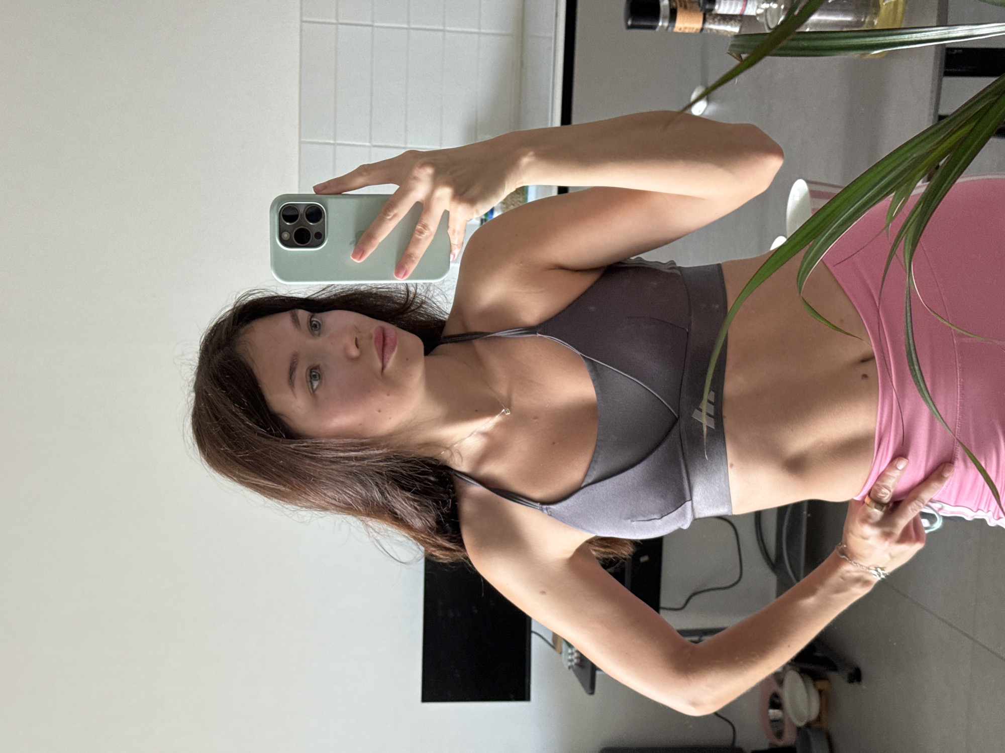 A young woman takes a selfie in a reflective surface. She is holding a light green smartphone with a multi-lens camera. She has brown hair, fair skin, and is wearing a gray sports bra and pink leggings. A silver necklace is visible around her neck. Her pose shows her right arm extended, holding the phone, while her left hand is near her hip, displaying rings and a bracelet. Part of a potted plant with long green leaves is in the foreground. The background includes a white tiled wall and a partial view of a dark-colored television screen or monitor on a desk. The environment appears to be an interior setting, potentially a room in a home or apartment. The precise location within Tel-Aviv, Israel cannot be determined from the image alone.