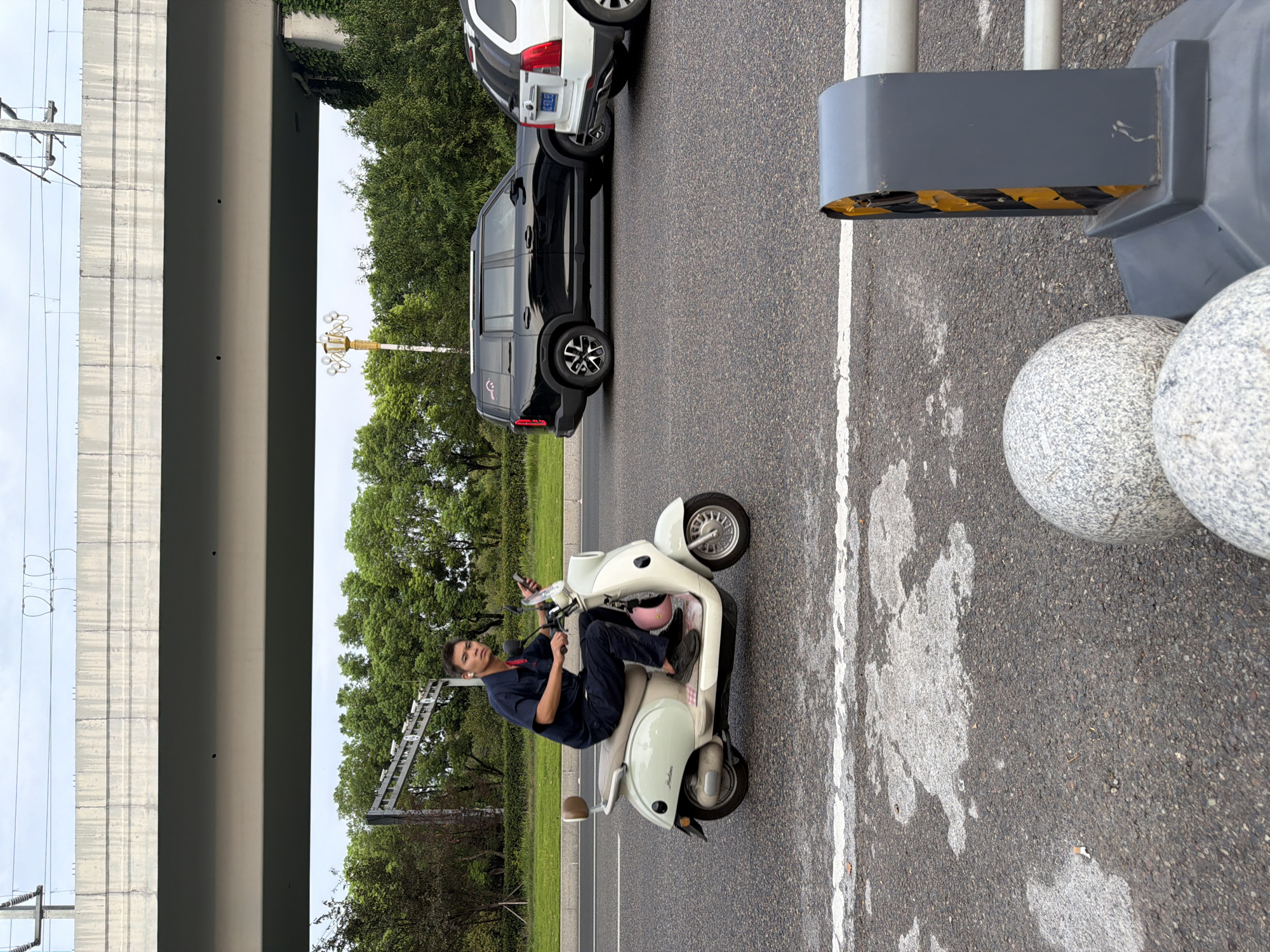 In Jiangjin, China, a man rides a light-colored scooter on a paved road with white lane markings. He is wearing a dark blue uniform with a red collar and has white earbuds in his ears. He is holding a mobile phone in his right hand. Behind him, a black SUV and a white SUV are driving in the same direction. The road is bordered by trees and a concrete overpass. In the foreground, there are two spherical granite bollards and a gray traffic barrier with yellow and black stripes. The sky is overcast.
