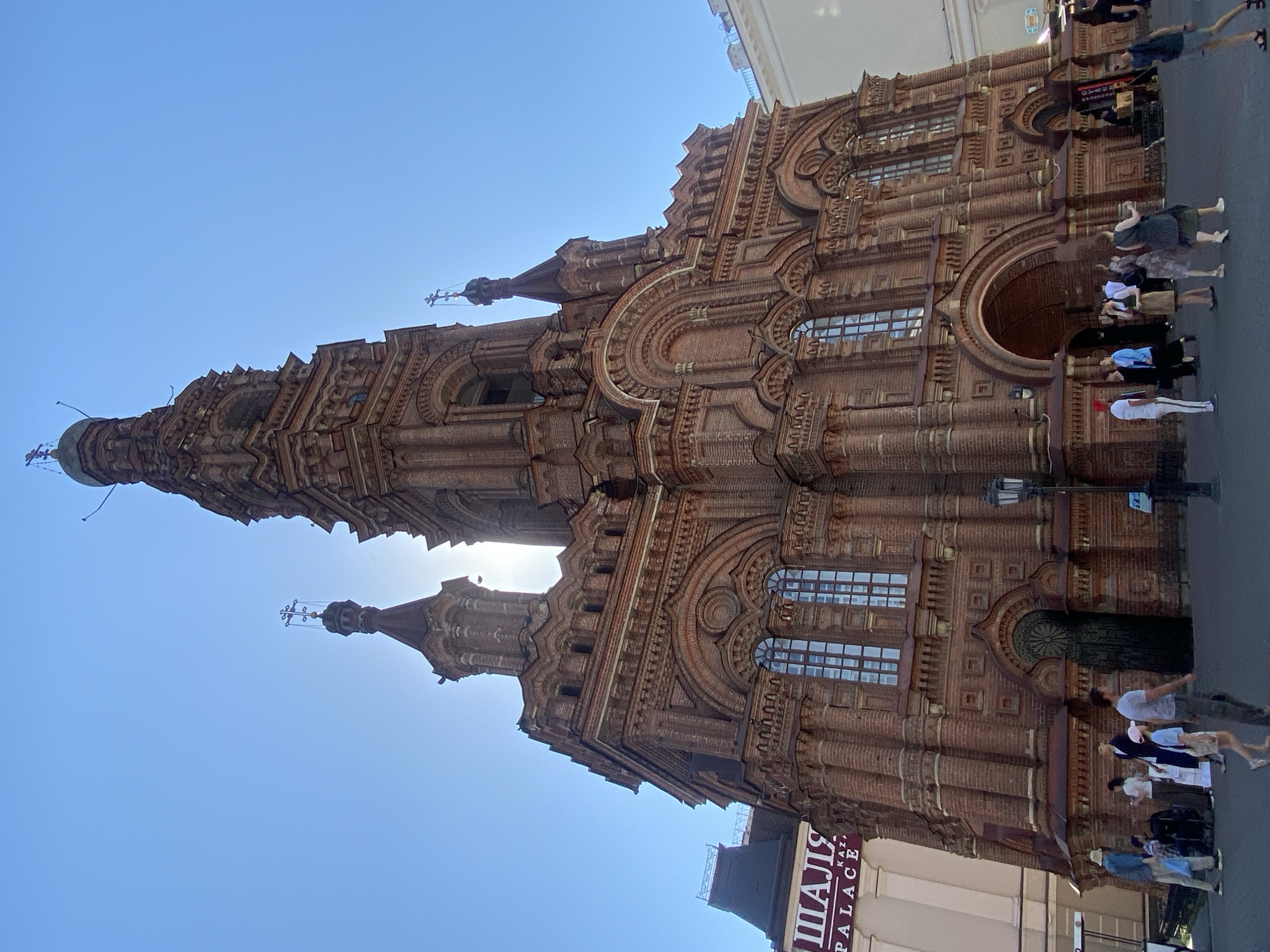 The image shows the Cathedral of the Annunciation in Kazan, Russia, viewed from a low angle. The cathedral is a multi-tiered, brick building with ornate architectural details including arched windows, decorative brickwork, and multiple small domes topped with crosses. The sun is partially obscured by the building, creating a bright spot in the sky. Several people are visible on the sidewalk in front of the cathedral. A building with signage partially visible is near the bottom of the frame. The sky is clear and blue.