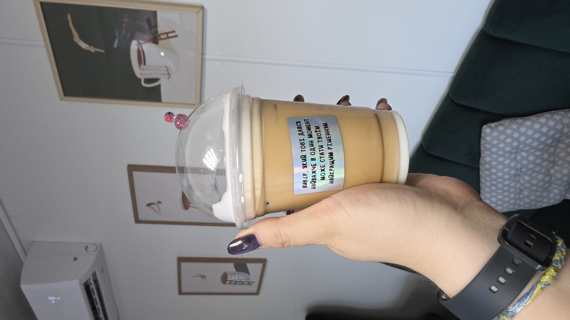 A close-up shot shows a hand holding a clear plastic cup filled with a light brown beverage and ice, topped with white foam. A holographic sticker with Ukrainian text is affixed to the cup. The background includes framed artwork on a white wall, and a portion of a dark green upholstered couch. The scene appears to be indoors, possibly in a cafe or public space. The air conditioner unit is visible on the wall. The overall impression is casual and everyday. There is no discernible indication of the city of Kyiv, Ukraine.