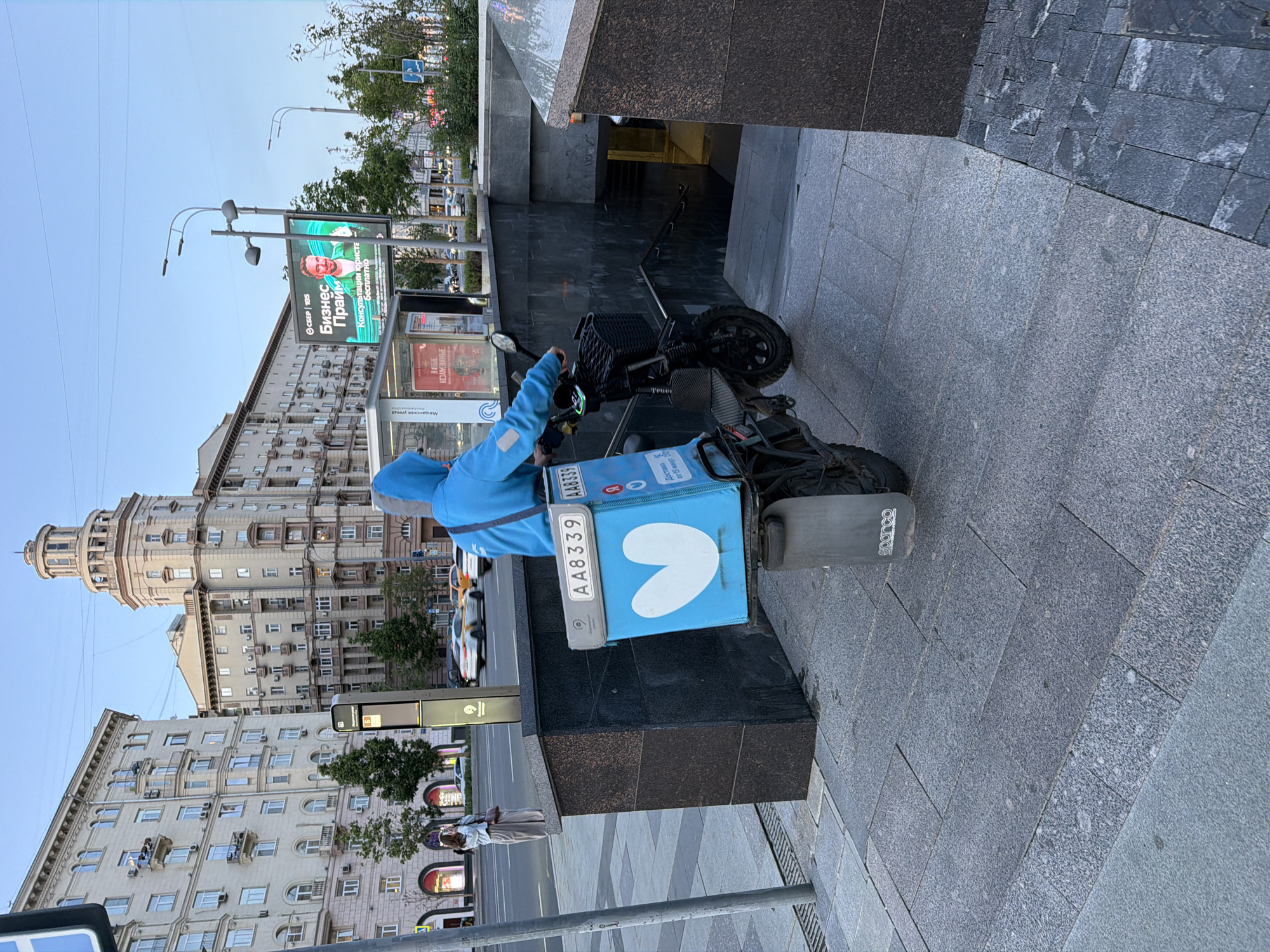 An outdoor, vertical-format photograph captures a vibrant urban scene in Moscow, Russia, under clear daylight. The central subject is a delivery person, clad in a light blue hooded jacket, paused on an electric bicycle or scooter. A prominent, bright blue delivery box, featuring a white heart-shaped logo and the alphanumeric code "AA8339" repeated twice, is affixed to the rear of the vehicle. The box also displays a distinct "Y" logo with Russian text reading "Доставка от 15 минут" (Delivery from 15 minutes). The mudguard of the bike clearly shows the "Sparco" brand.
The delivery person is positioned on a paved sidewalk made of grey granite tiles, adjacent to a dark, granite-clad structure that serves as a metro station entrance, with stairs descending into it. Beyond this structure, a street with visible cars and green trees can be seen.
In the background, tall, multi-story buildings of a classic architectural style, light in color, line the street. These buildings are characterized by numerous windows and many external air conditioning units; one building features a distinctive, rounded tower at its top. A large digital billboard is visible on the right, displaying a smiling man and Russian text that reads "Бизнес Прайм" (Business Prime), "СБЕР | EBS", and "Консультации юриста бесплатно" (Lawyer consultation free). Other smaller signs with Russian text such as "Мещанская улица" (Meshchanskaya Street) and "НОВЫЕ НЕЗАМЕНИМЫЕ" (NEW IRREPLACEABLE) are also faintly visible. The sky is clear and light blue, indicating fair weather. The scene portrays a typical bustling city environment.
